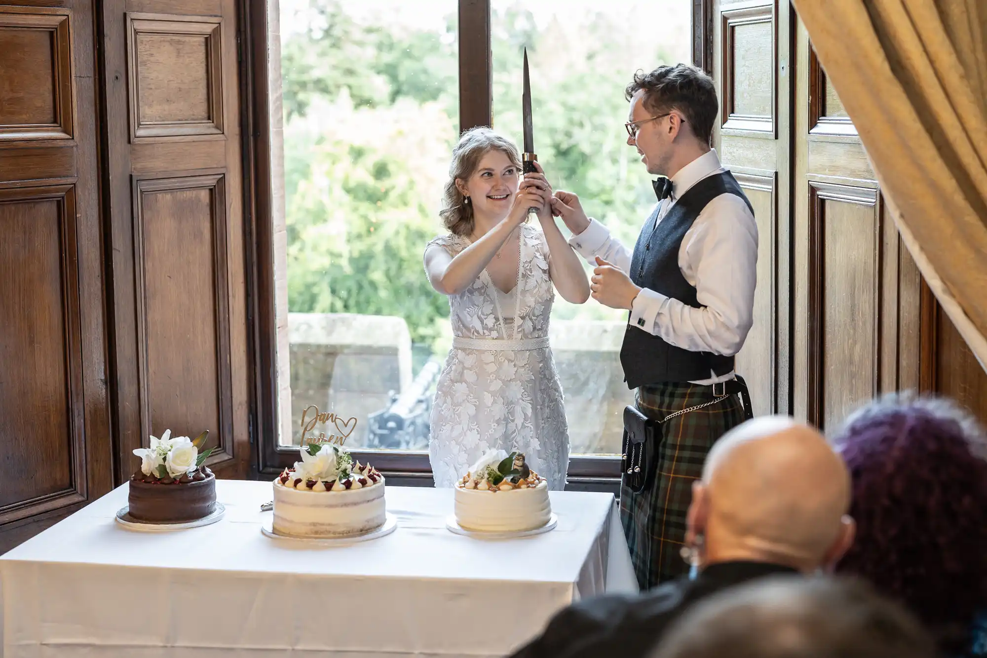 A couple stands by a table with three cakes, holding a knife together, presumably to cut one of the cakes. The background features wooden paneling and a window showing greenery outside.