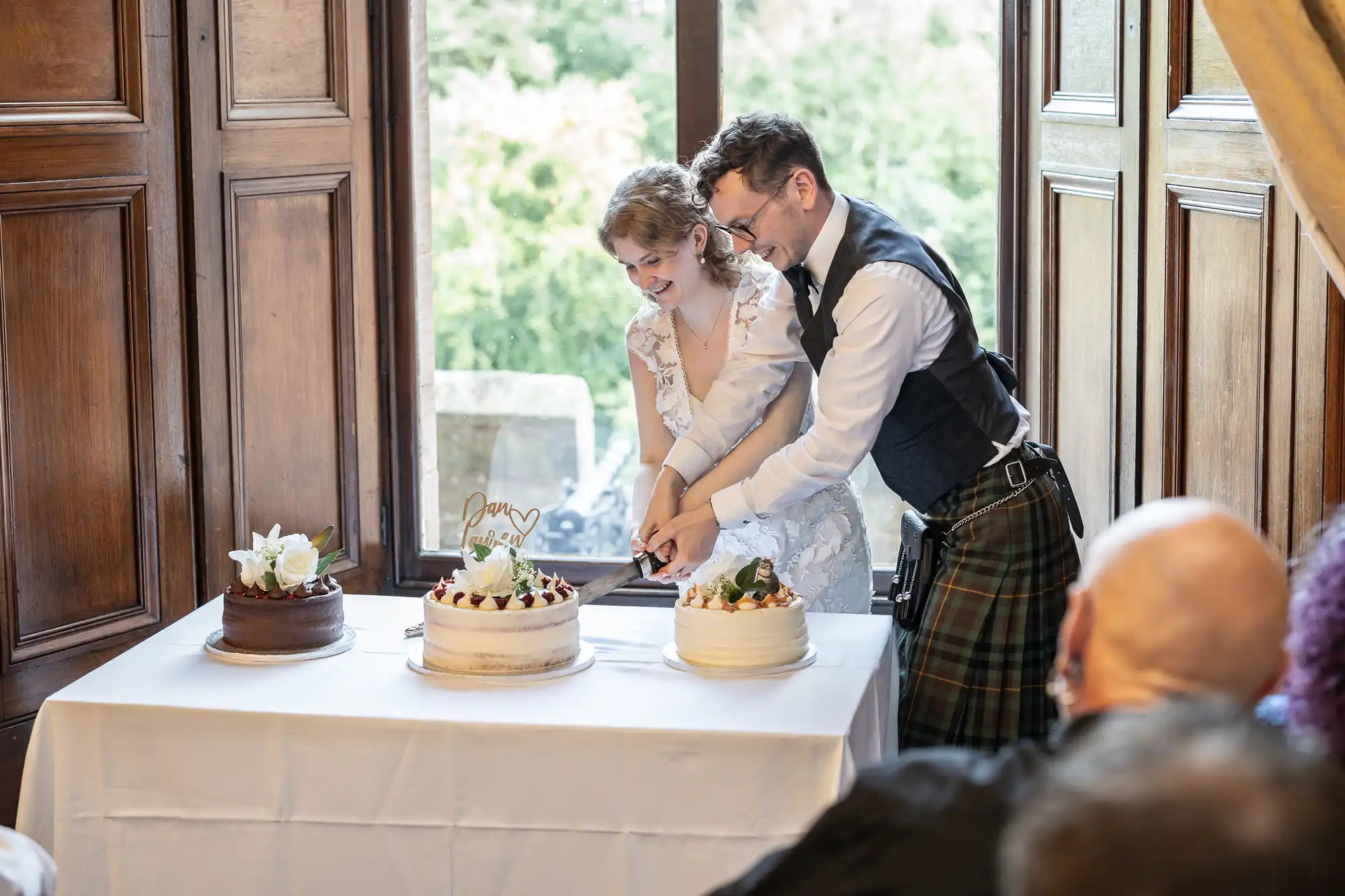 A couple is cutting a white-frosted cake together at a celebration. They stand behind a table with three different cakes on it. People are watching in the foreground.