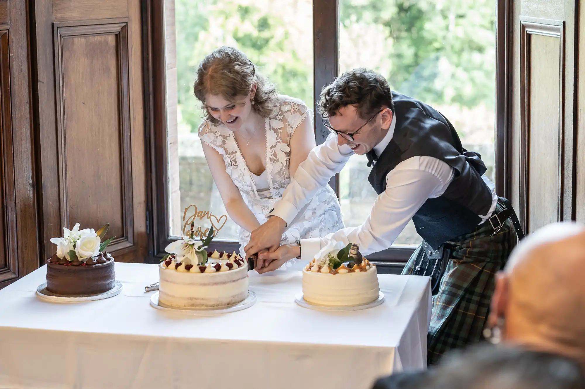 A couple, dressed in wedding attire, cuts a cake together at a table with three cakes. The man wears a kilt, and the woman wears a white wedding dress.