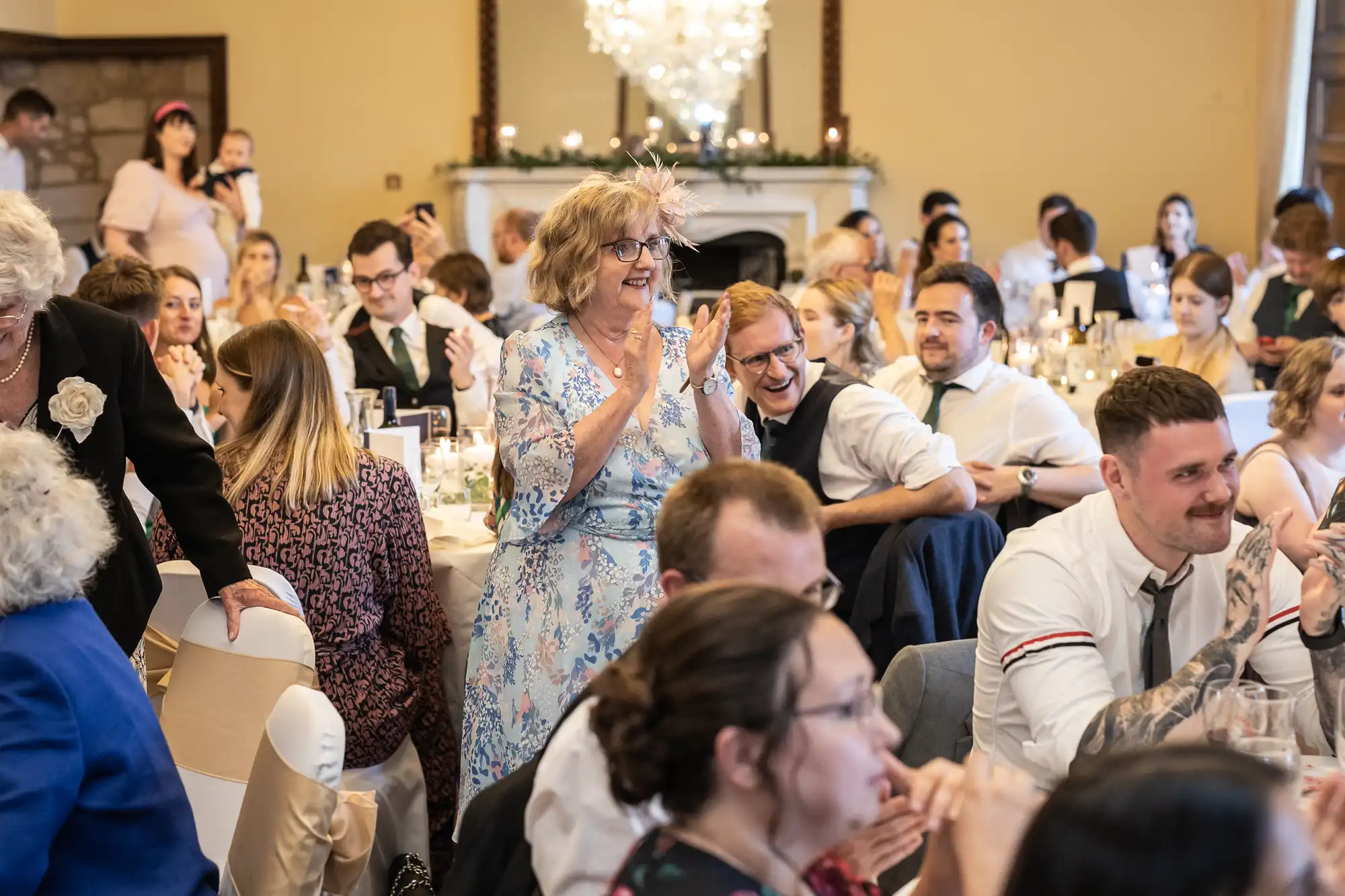 A group of people are seated at a formal event. A woman in a floral dress is standing and clapping while others are seated around her, engaged in conversation and dining.