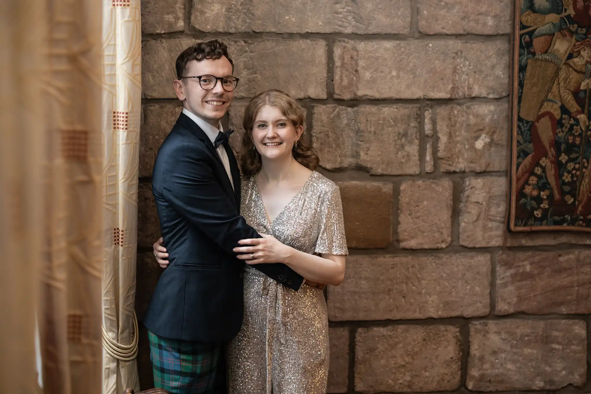 A man in a dark suit and glasses stands with a woman in a sparkly dress, both smiling and posing in front of a brick wall indoors.