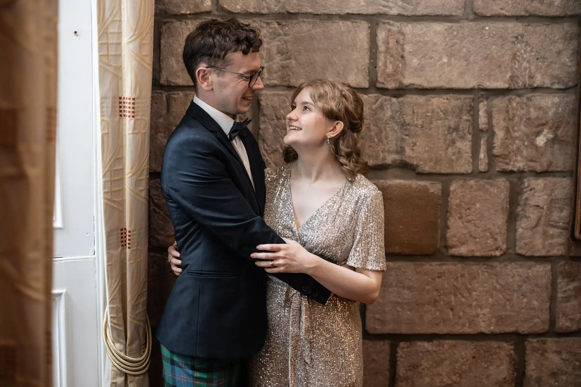 A man in a dark suit and a woman in a glittery dress stand in front of a stone wall, smiling and holding each other.