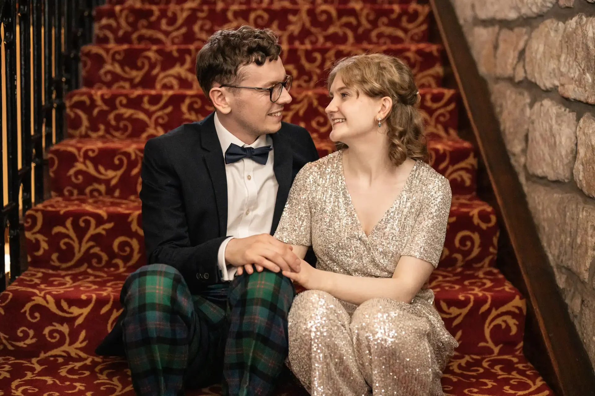 A couple dressed formally sits on a patterned carpeted staircase, facing each other and holding hands, appearing happy and engaged in conversation.
