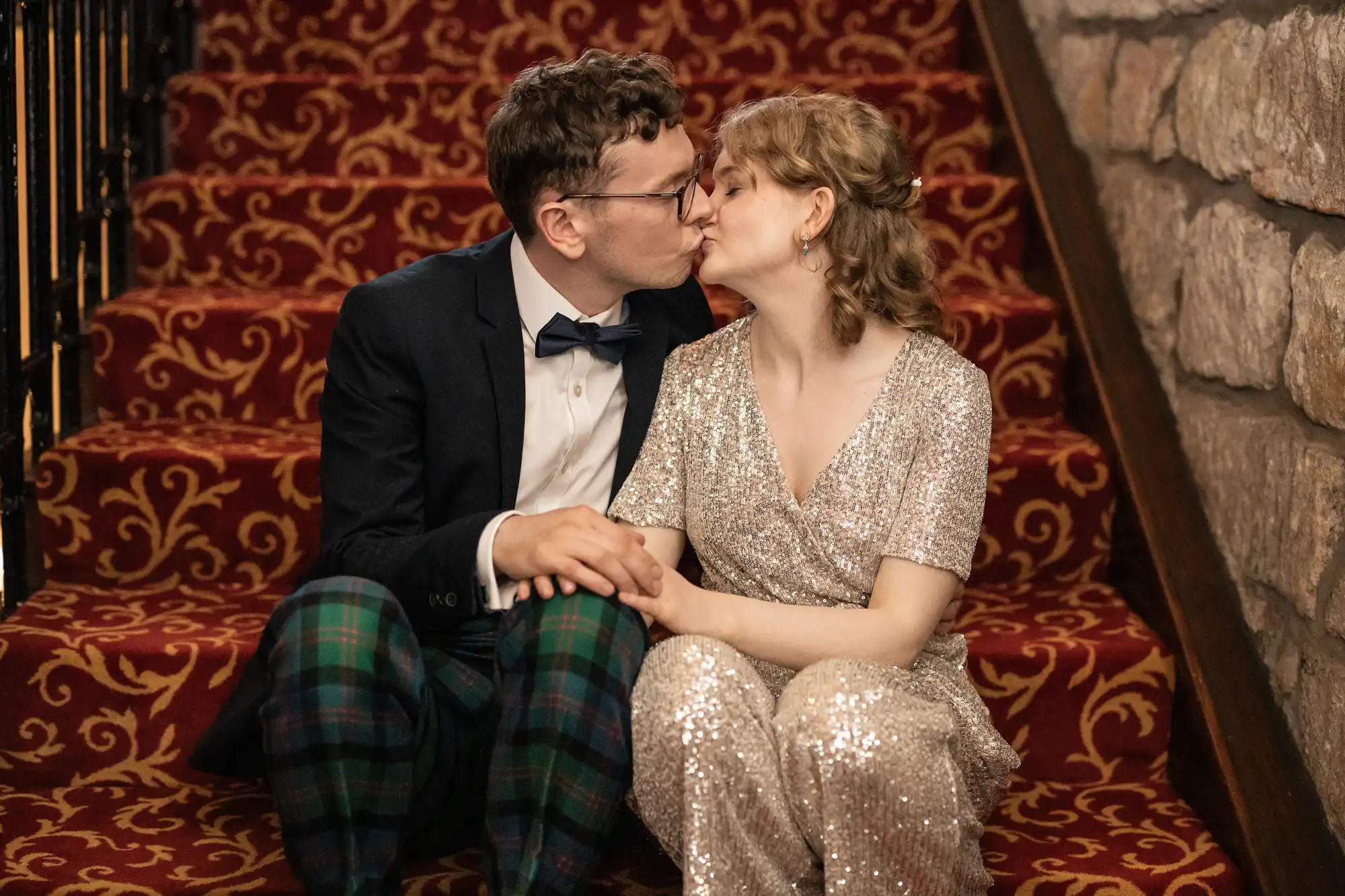 A couple, dressed formally, sits on red patterned stairs, holding hands and kissing. The man wears a blazer with plaid pants, and the woman wears a sequined dress.