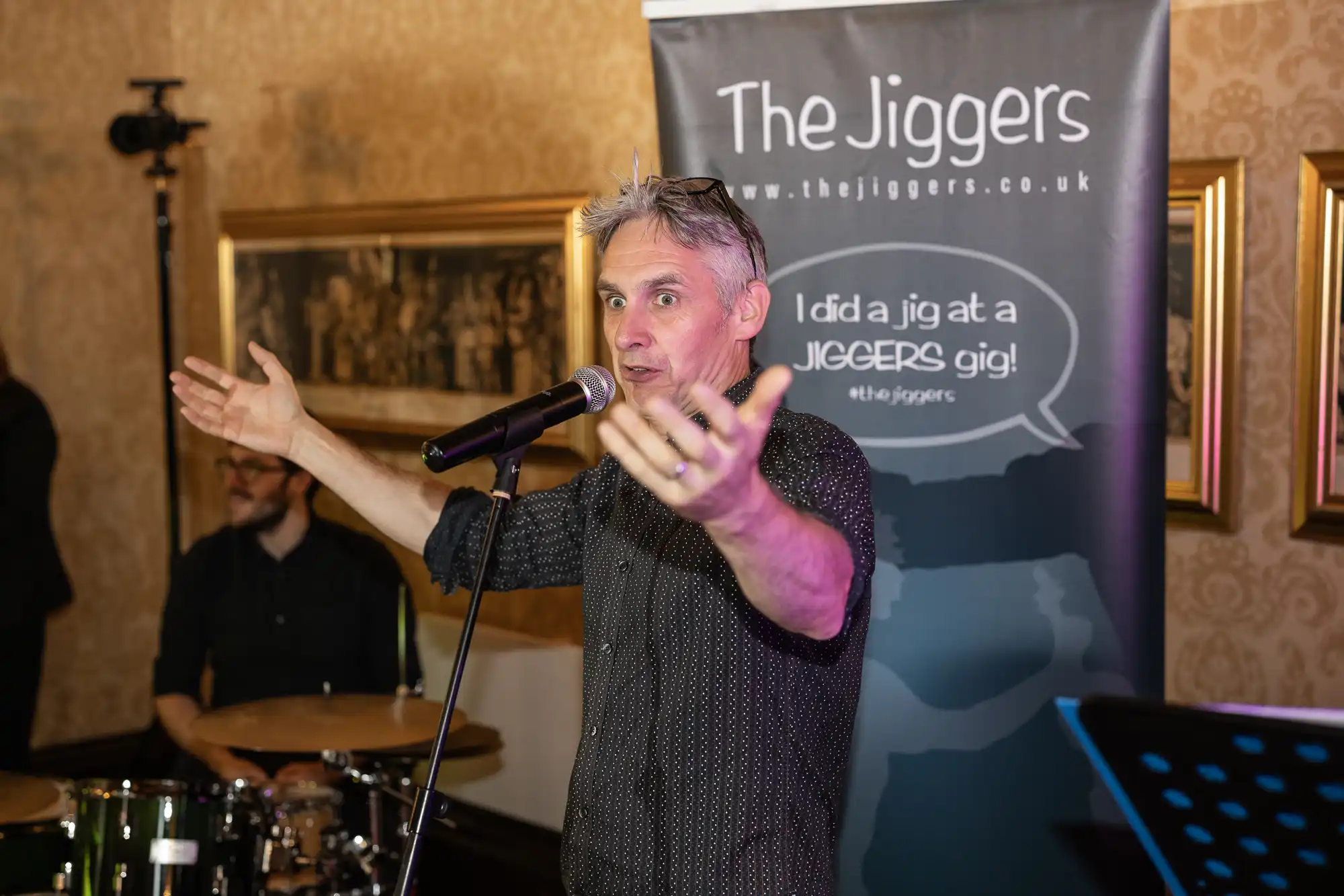 A man with gray hair speaks into a microphone with his arms outstretched. Behind him is a banner that says "The Jiggers" and "I did a jig at a JIGGERS gig!" Drummer is partially visible in the background.