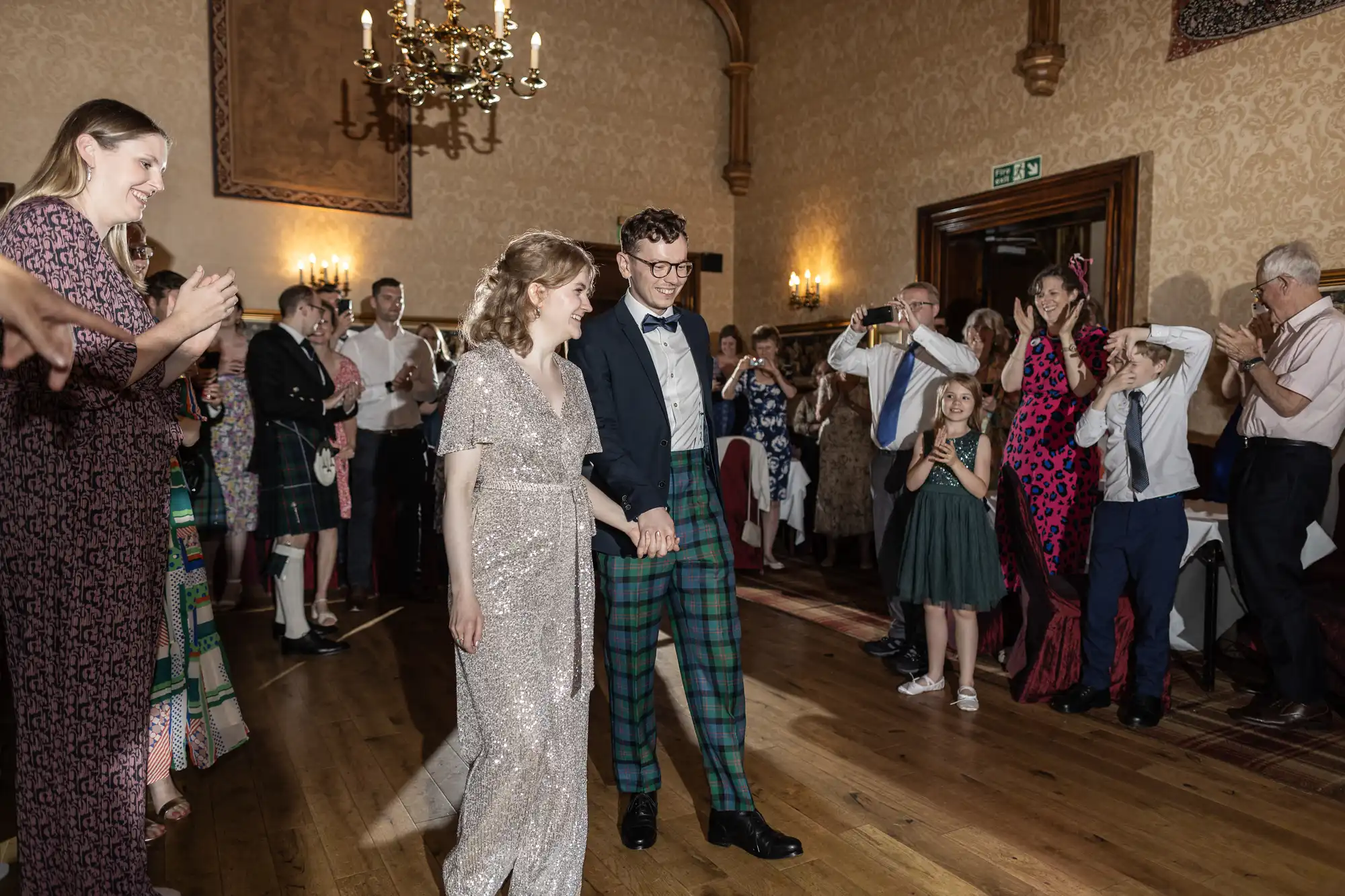 A couple, dressed formally, walks hand in hand on a wooden floor surrounded by clapping and smiling guests in a warmly lit room with ornate decor and a chandelier.