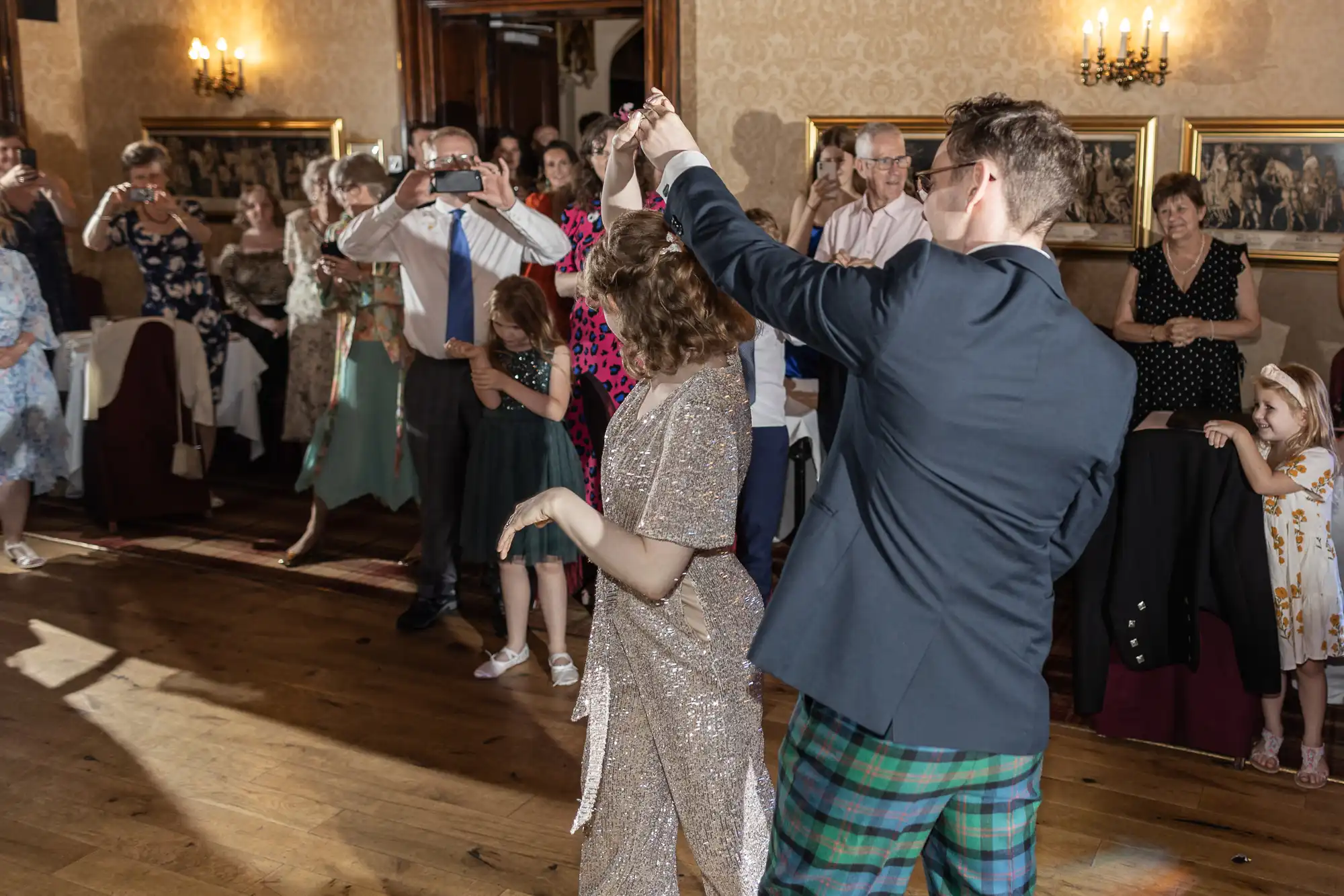 Two people dance in the center of a room while a group of onlookers, some taking photos, watch. The dancers are wearing formal attire; the person on the right is in a suit jacket and tartan trousers.