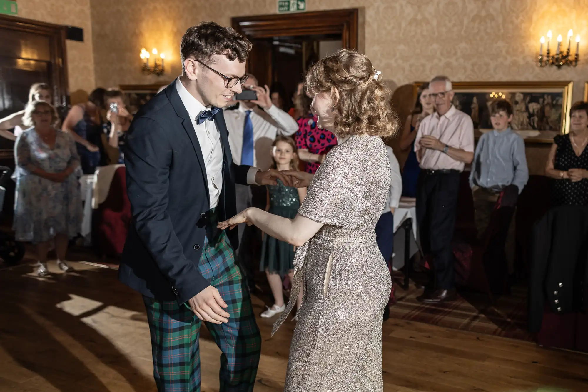 A man and a woman dance together at a formal event while guests look on. The man wears a suit with plaid pants, and the woman wears a sparkly dress. The room is decorated with ornate wallpaper and chandeliers.