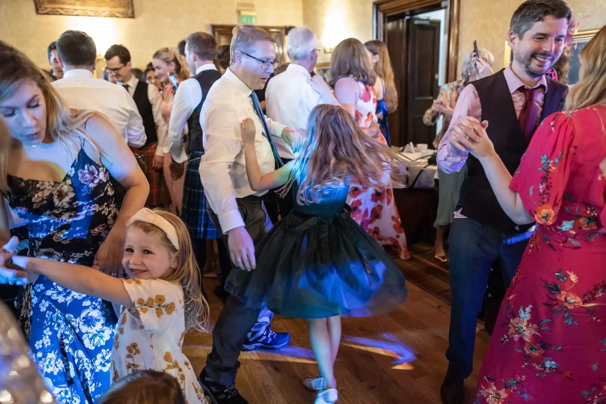 A group of people, including children, dancing and socializing in a lively indoor setting. The atmosphere is festive, with attendees dressed in formal and semi-formal attire.