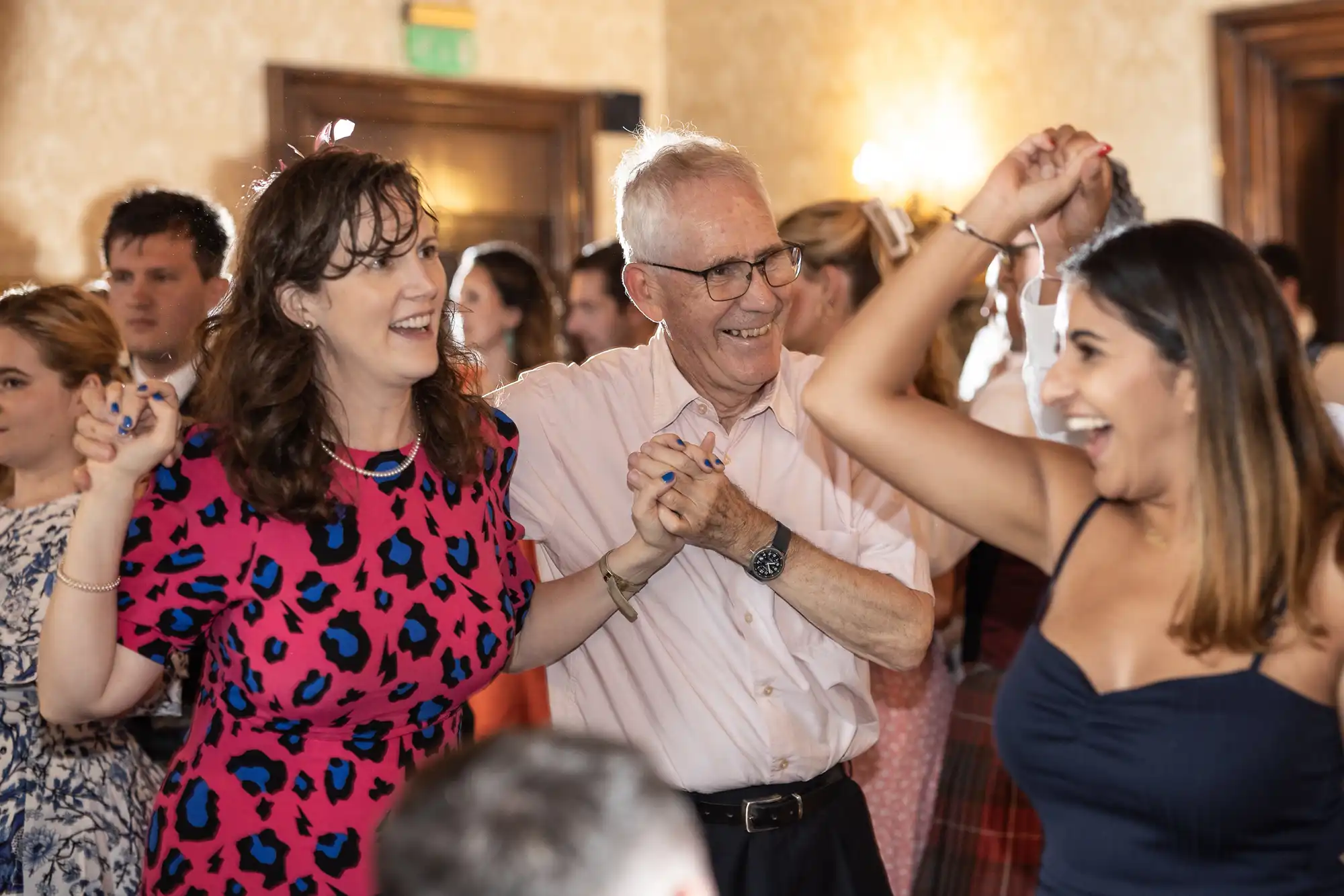 A group of people smile and dance while holding hands at an indoor social event. The setting suggests a joyful atmosphere.