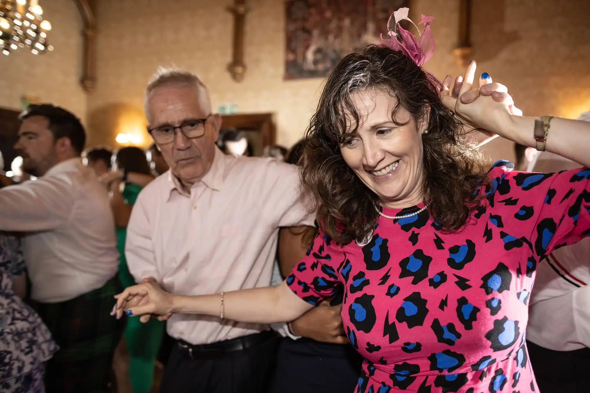 People dancing at a social event, a woman in a bright pink and blue dress in the foreground smiling and holding a pen, with other individuals in the background.