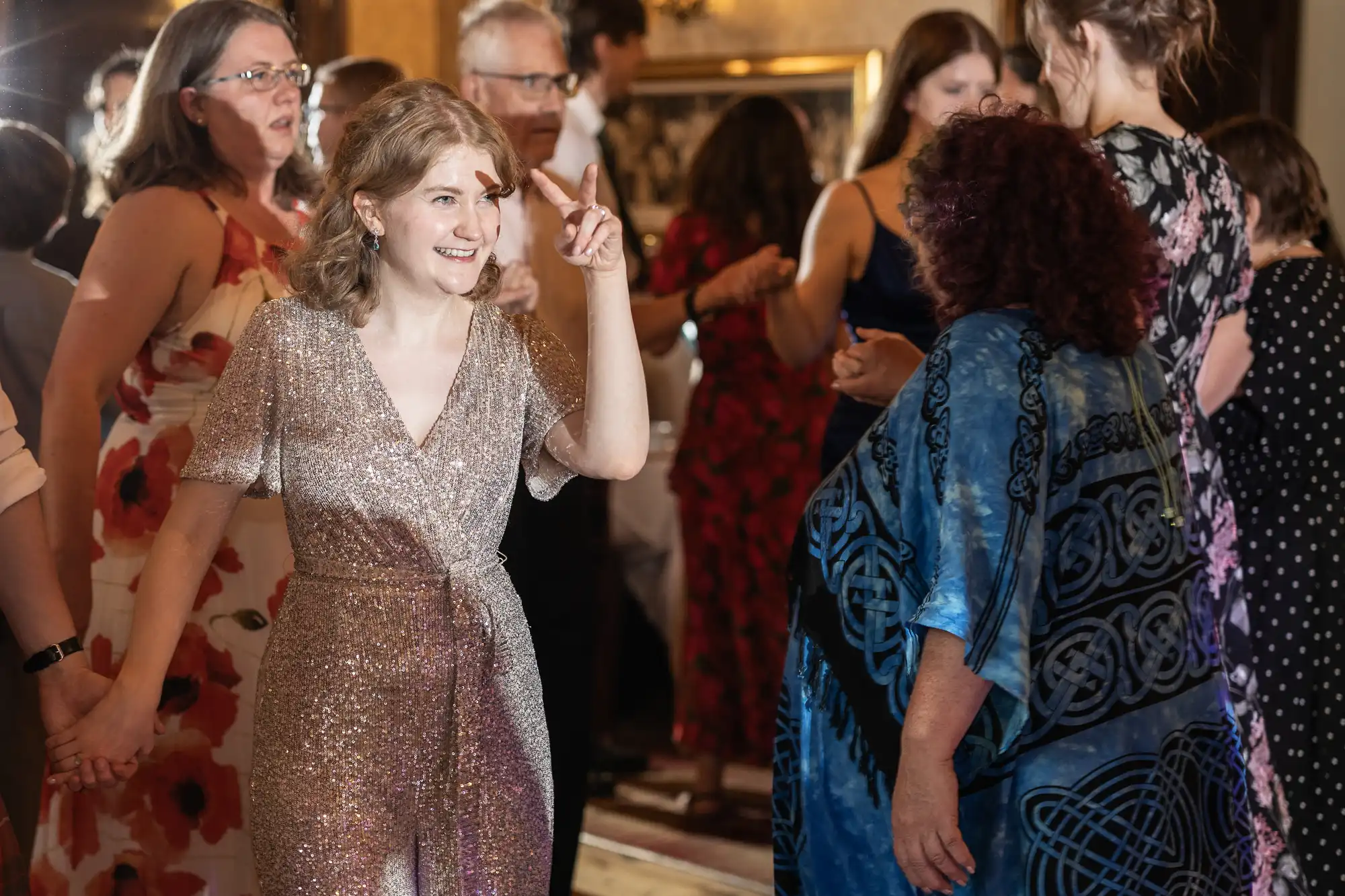 A woman in a sparkly dress smiles and gestures with her fingers while dancing in a crowded room. Other people are also engaged in conversation and dancing around her.