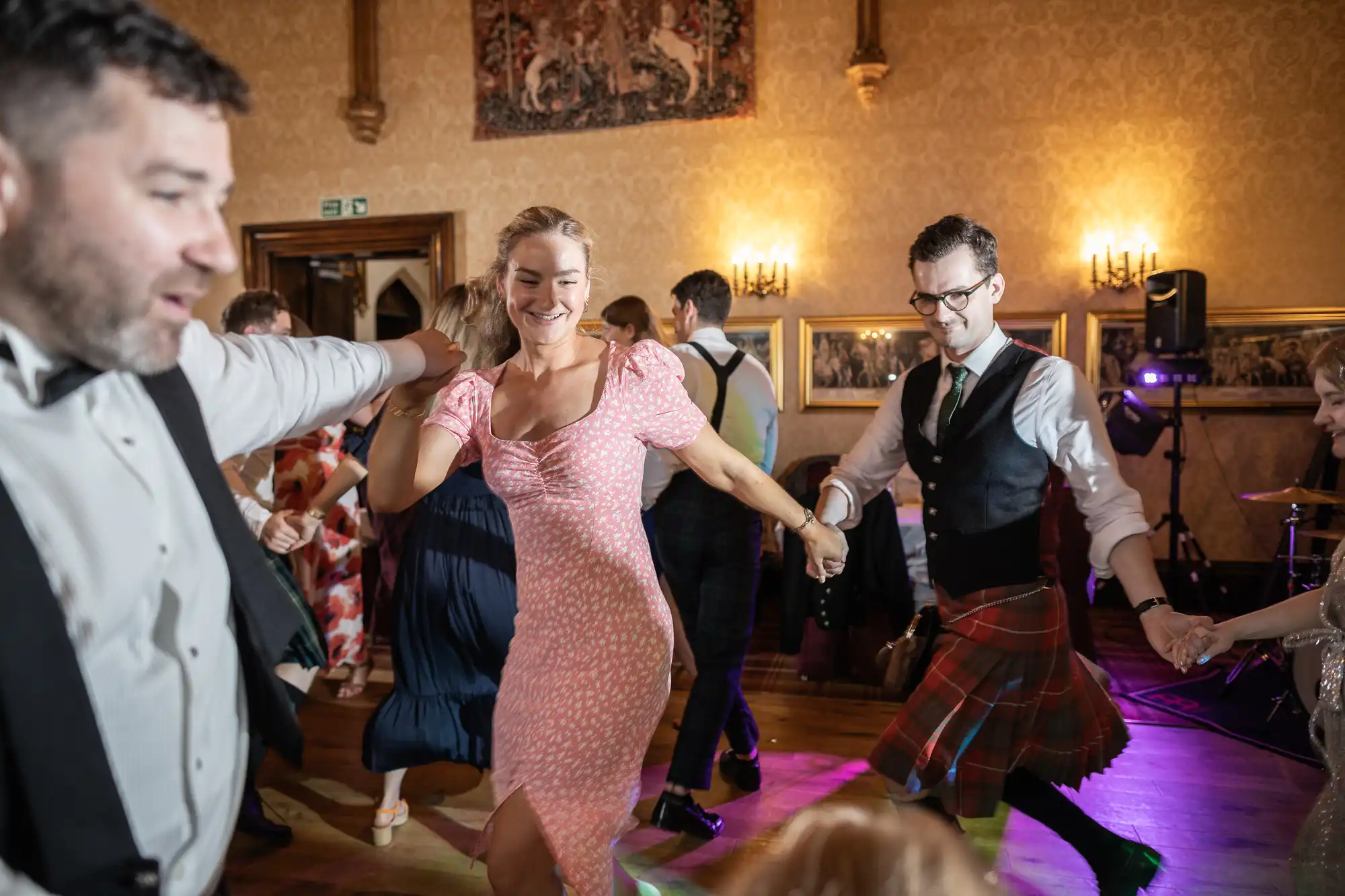 People dancing joyfully in a decorated hall, holding hands in a circle. Some are wearing formal attire, including kilts, while a few smile and face the camera.