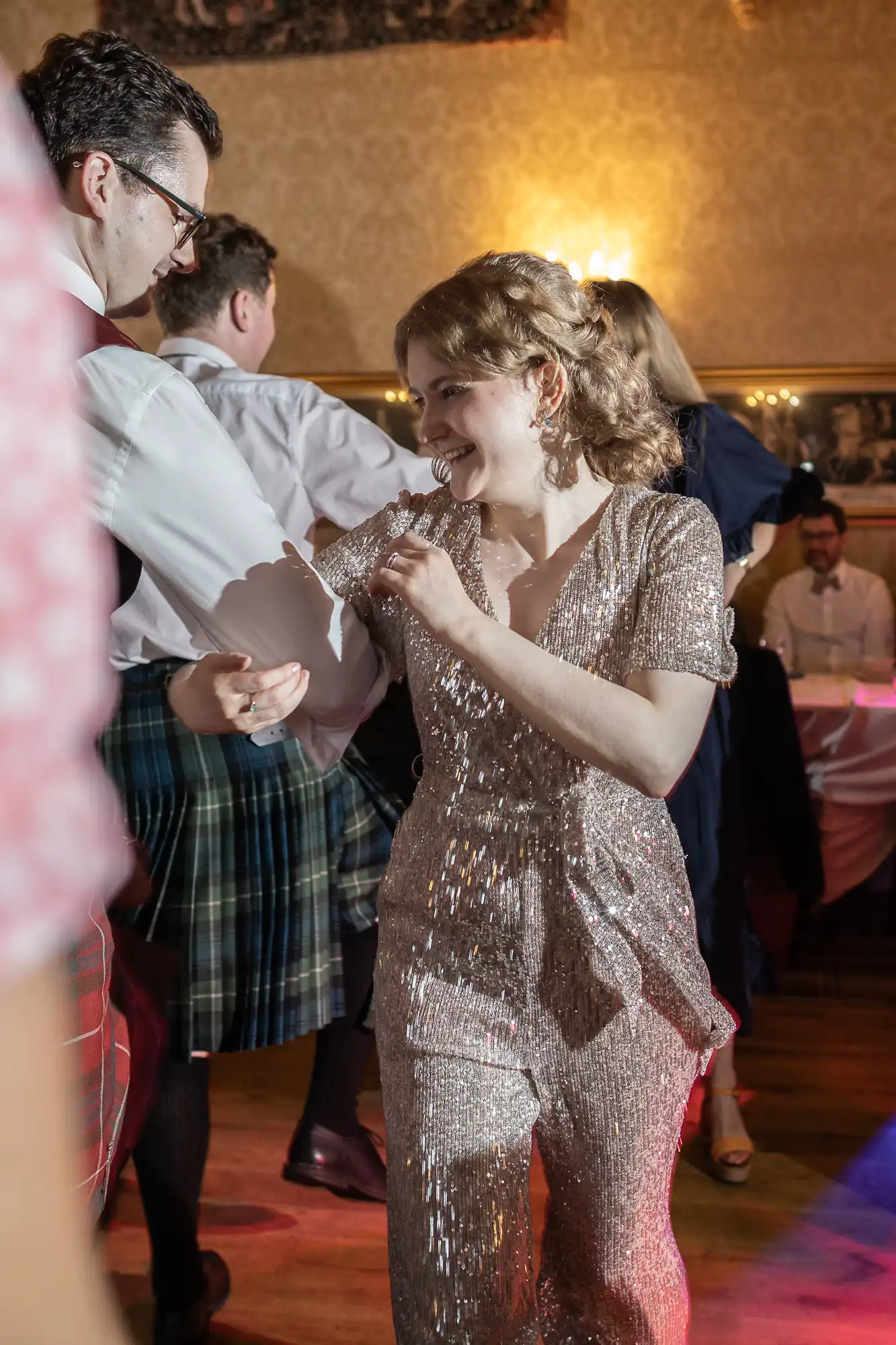 A woman in a glittering outfit dances with a man in a kilt at a lively event, other attendees can be seen in the background.