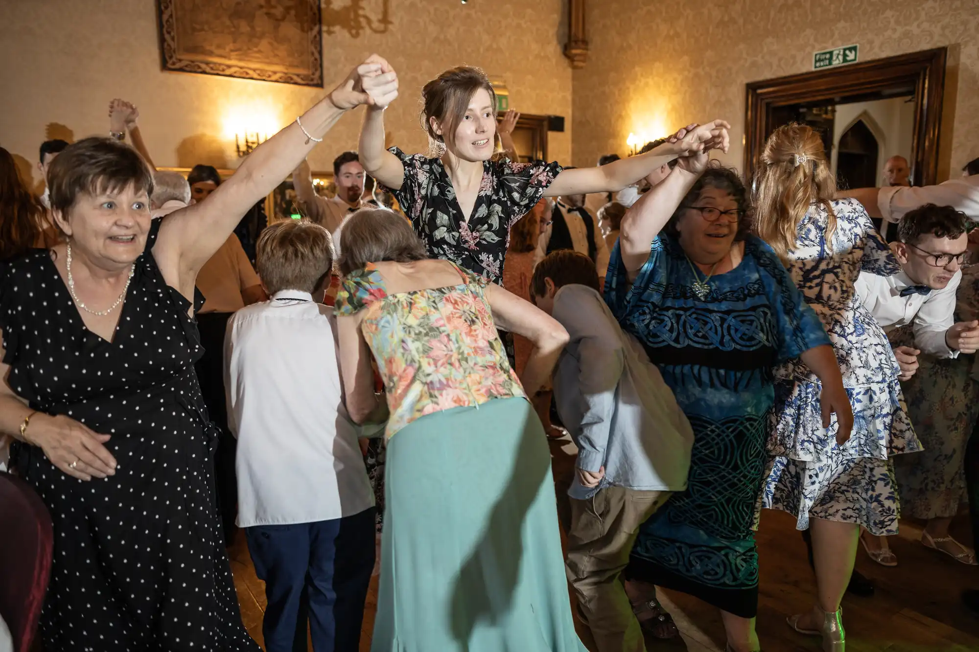 A group of people are dancing energetically in a room decorated with ornate wall hangings and dim lighting. Some individuals have their hands raised, and everyone appears to be enjoying the moment.