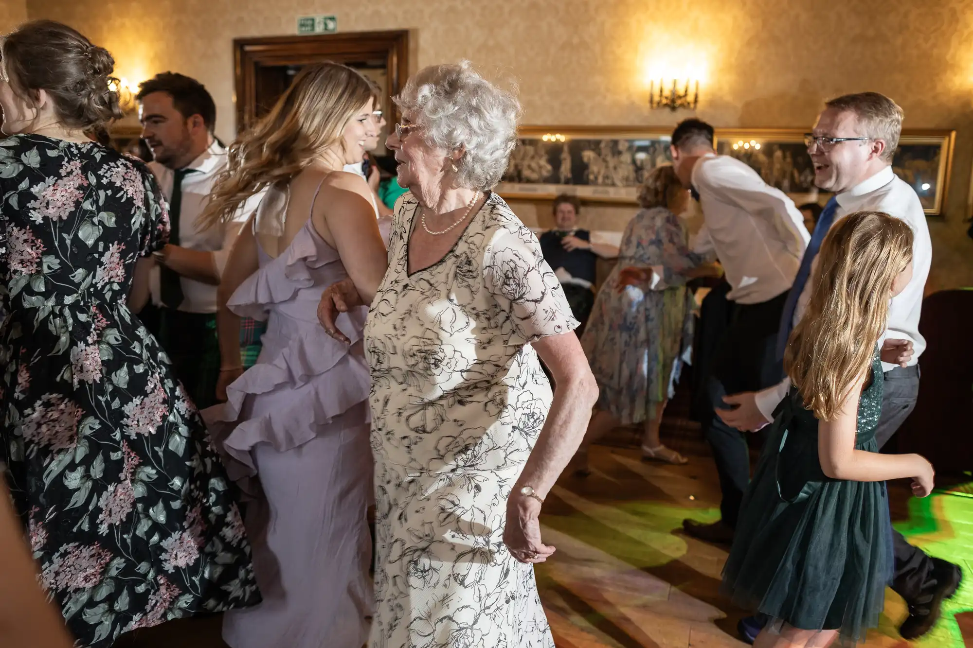 A group of people of varying ages dance joyfully in a warmly lit room with floral wallpaper and framed pictures on the wall.