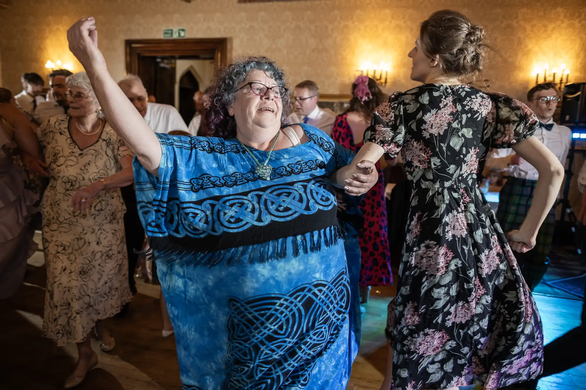 A group of people are dancing at a lively indoor event. A woman in a blue dress with intricate designs is holding hands and dancing with another woman in a floral dress.