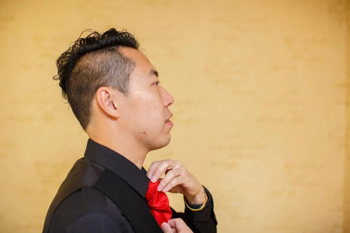 profile view as groom adjusts his red cravat in front of a mirror
