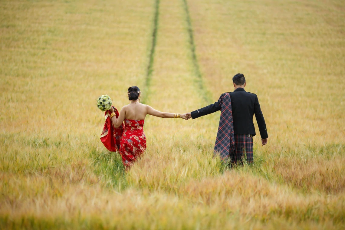Chinese bride wearing a red dress holds her husband's hand as they walk through a field