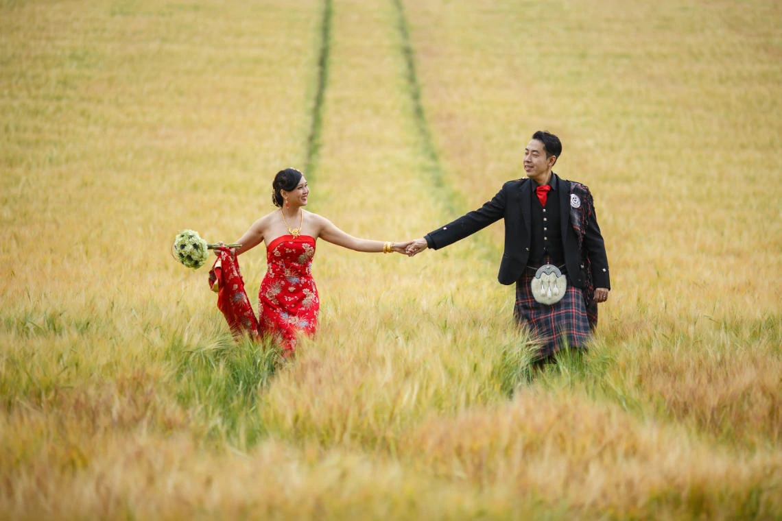 Chinese bride wearing a red dress holds her husband's hand as they walk through a field