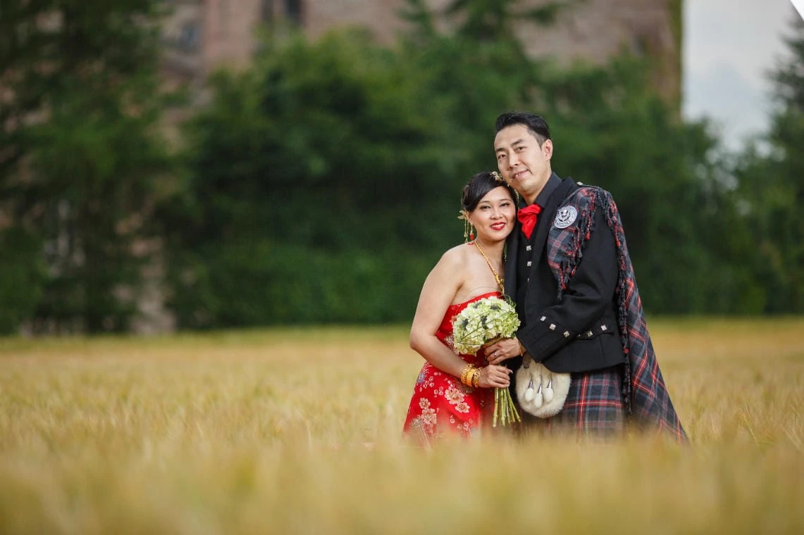 Chinese bride wearing a red dress embraces husband in the field next to the castle