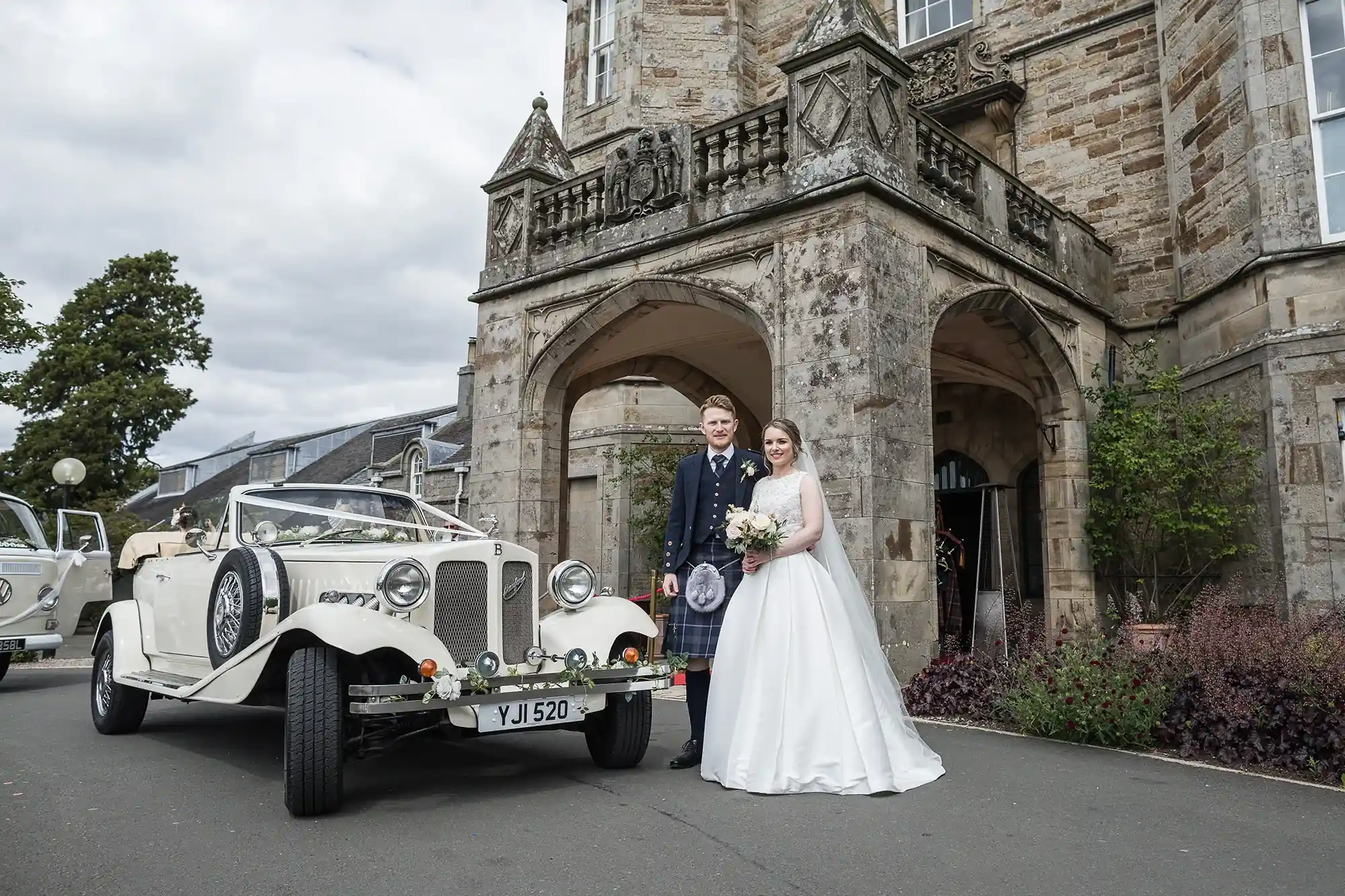 A newlywed couple posing in front of a vintage car at the entrance of an old stone building. the groom is wearing a kilt and the bride is in a white gown, holding a bouquet.