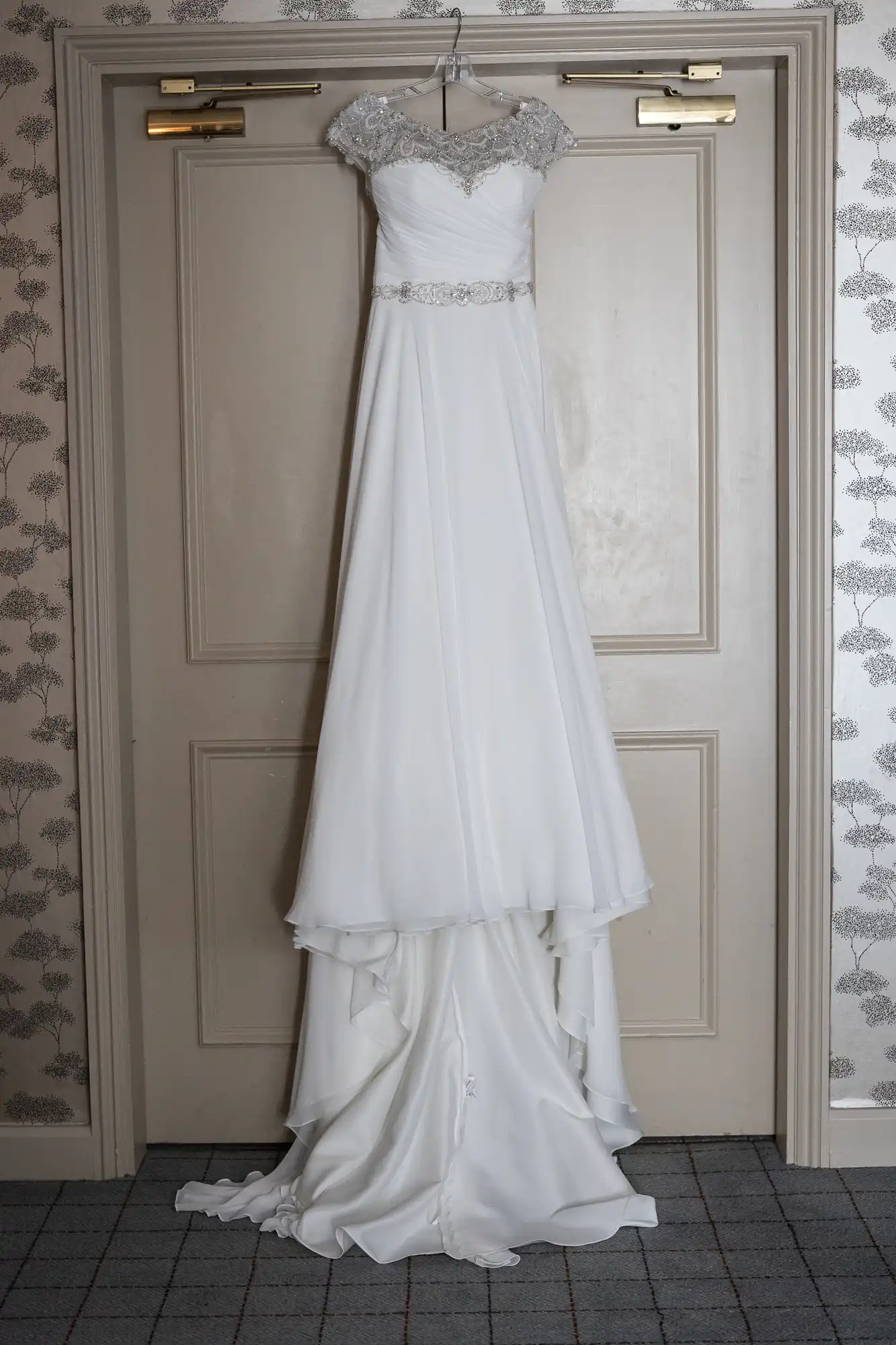 A white wedding dress with lace embellishments on the sleeves and neckline, hanging on a wooden door.