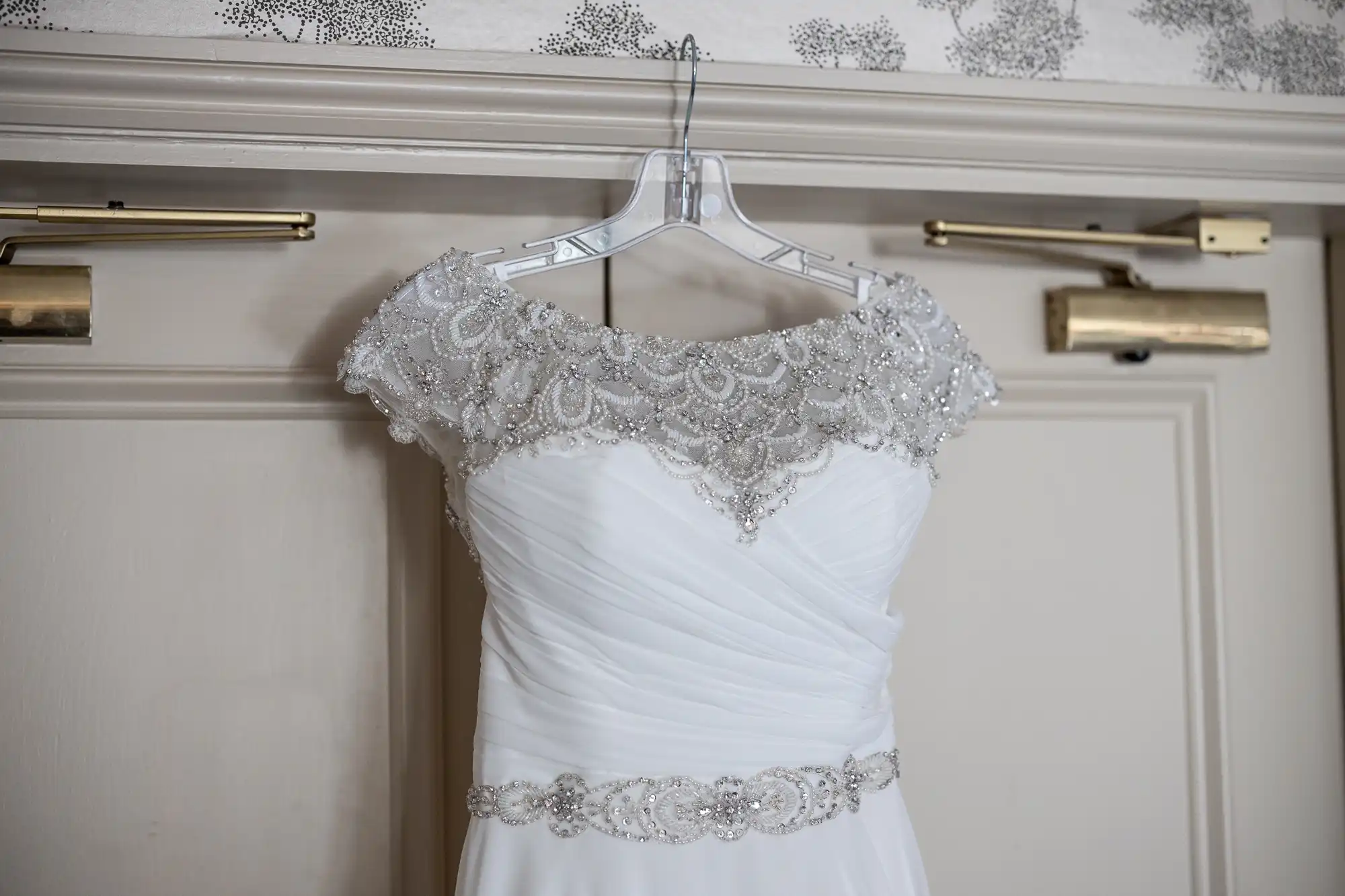 A white wedding dress with intricate beadwork on the neckline and waist is hanging on a clear plastic hanger against a beige door.