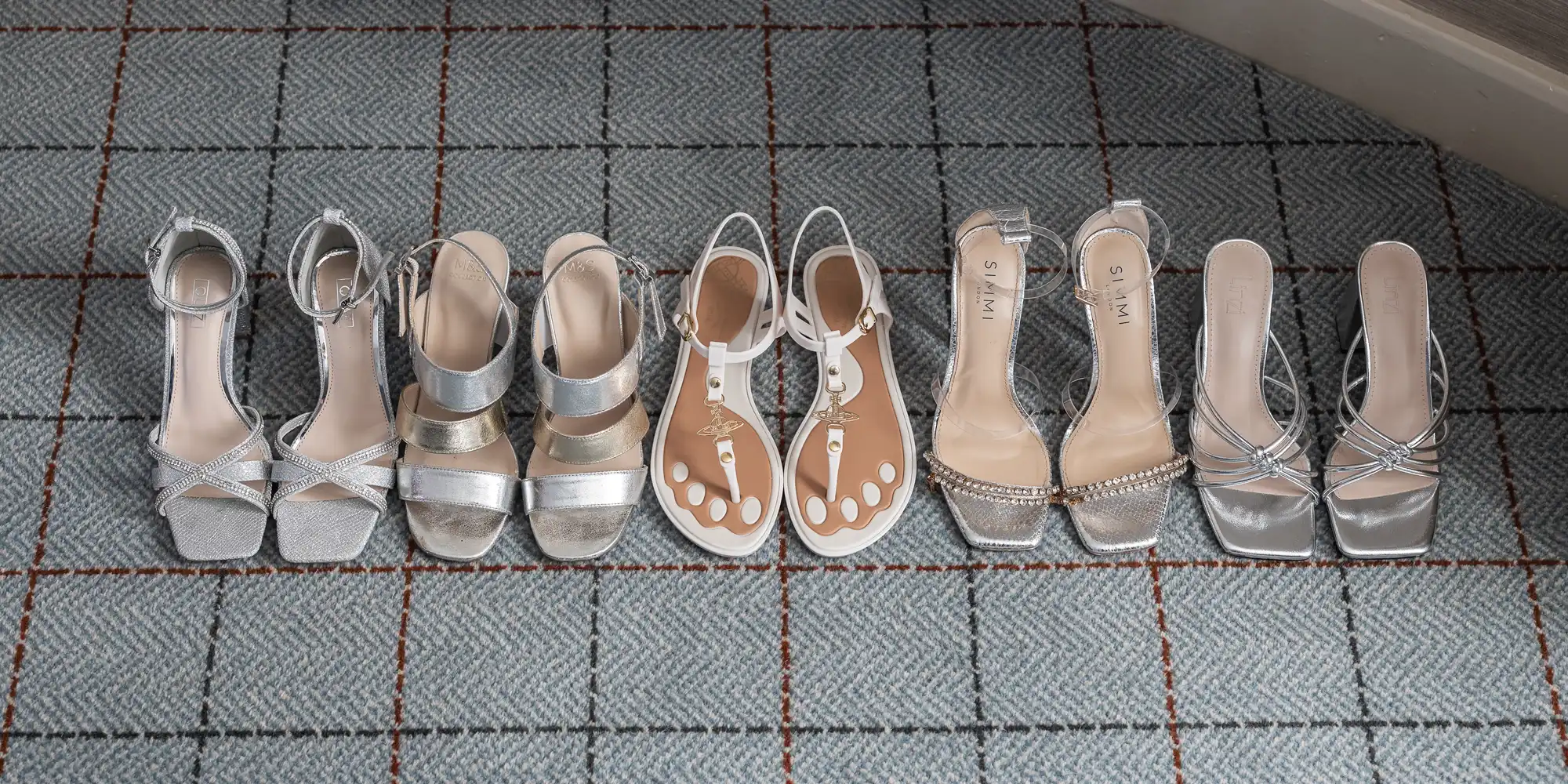 Four pairs of women's shoes are lined up side by side on a carpeted floor. The pairs consist of silver high heels and a single pair of flat sandals with white straps and clear toe dividers.