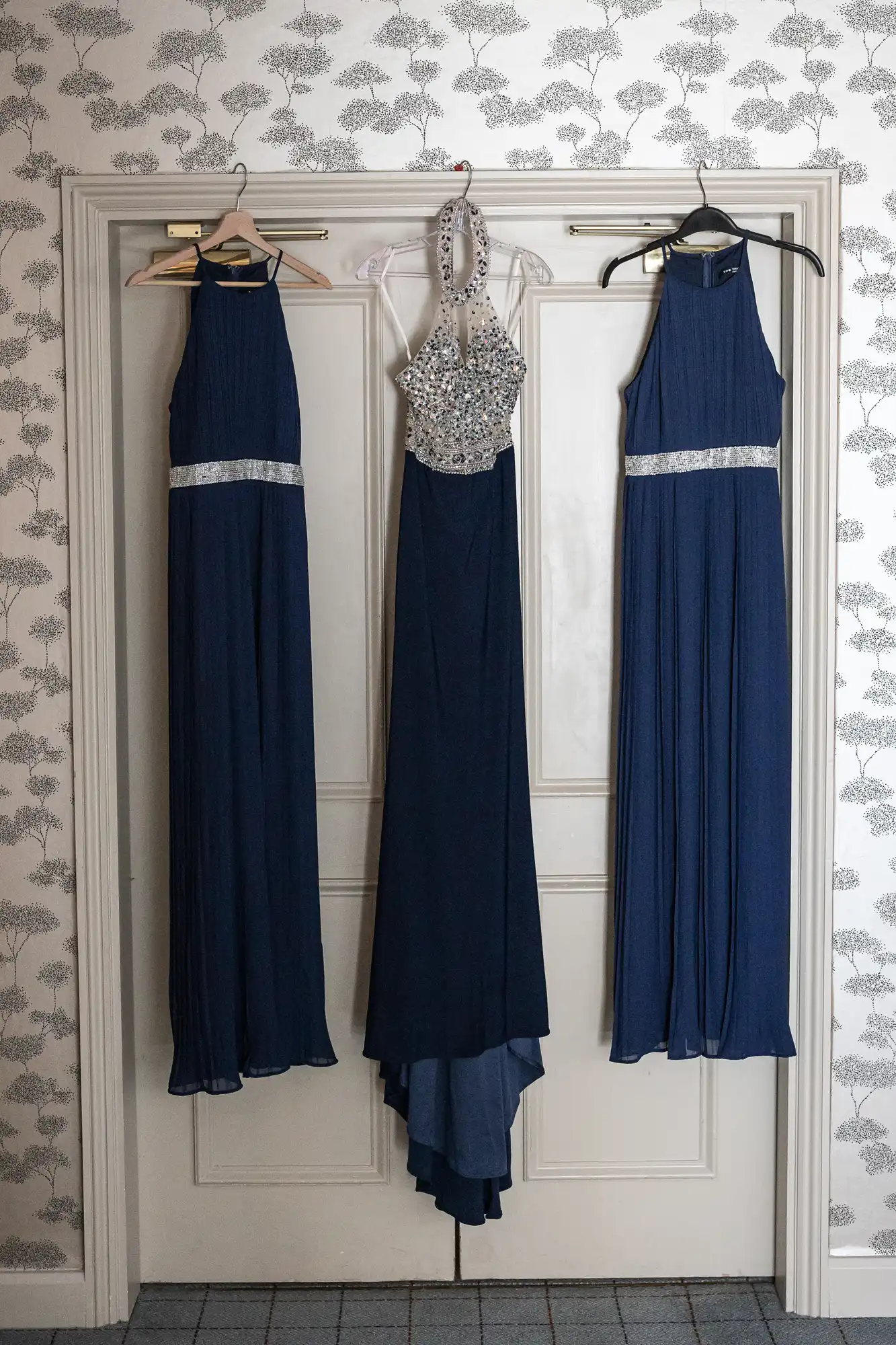 Three blue gowns with silver embellishments are hanging on a white door against a patterned wallpaper background.