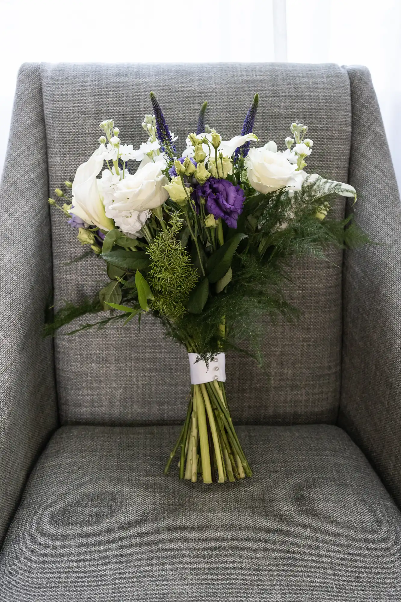 A bouquet of white and purple flowers with green foliage is placed upright on a gray upholstered chair.