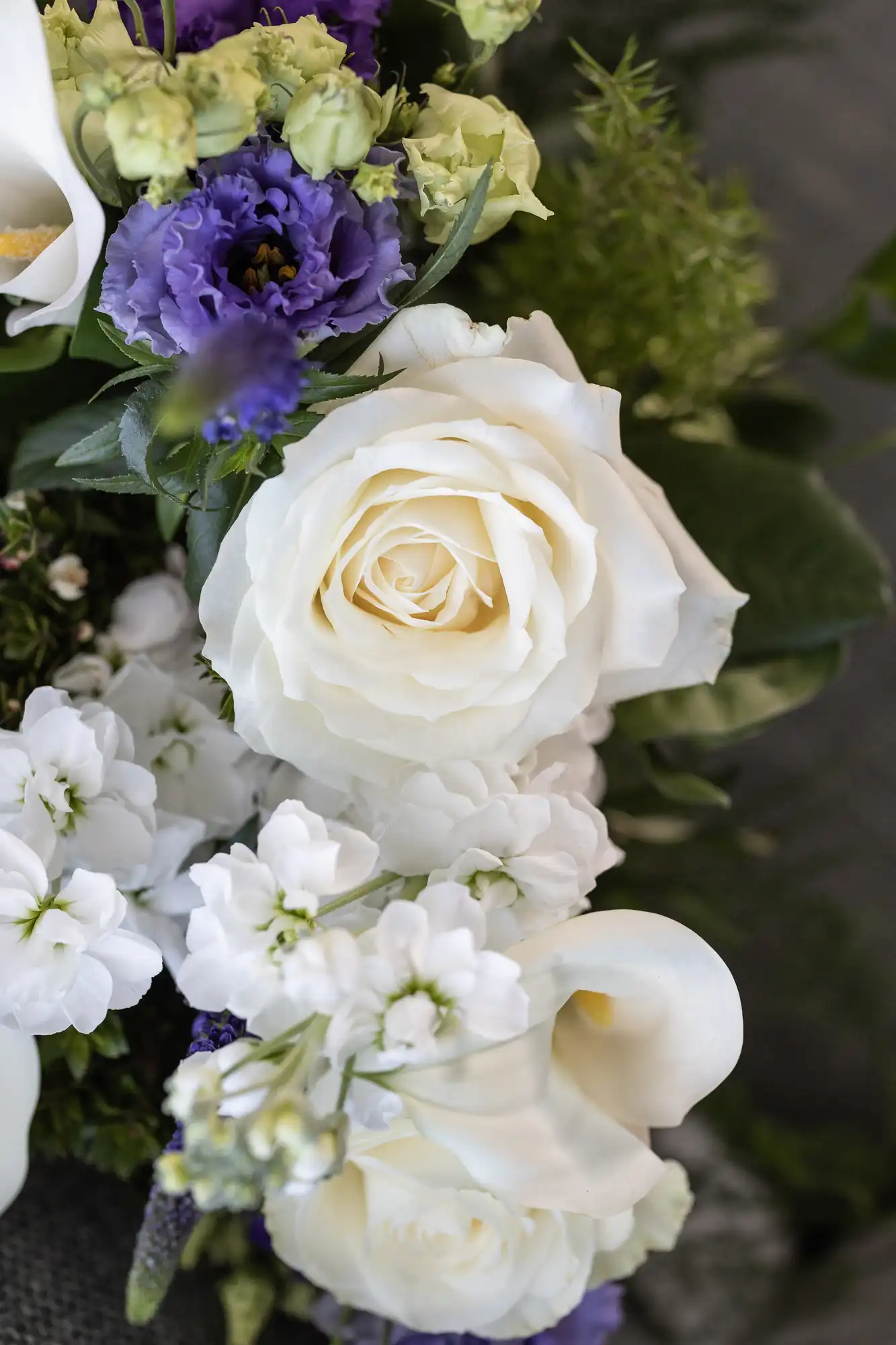 A close-up of a floral bouquet featuring a prominent white rose surrounded by purple lisianthus and other small white flowers.