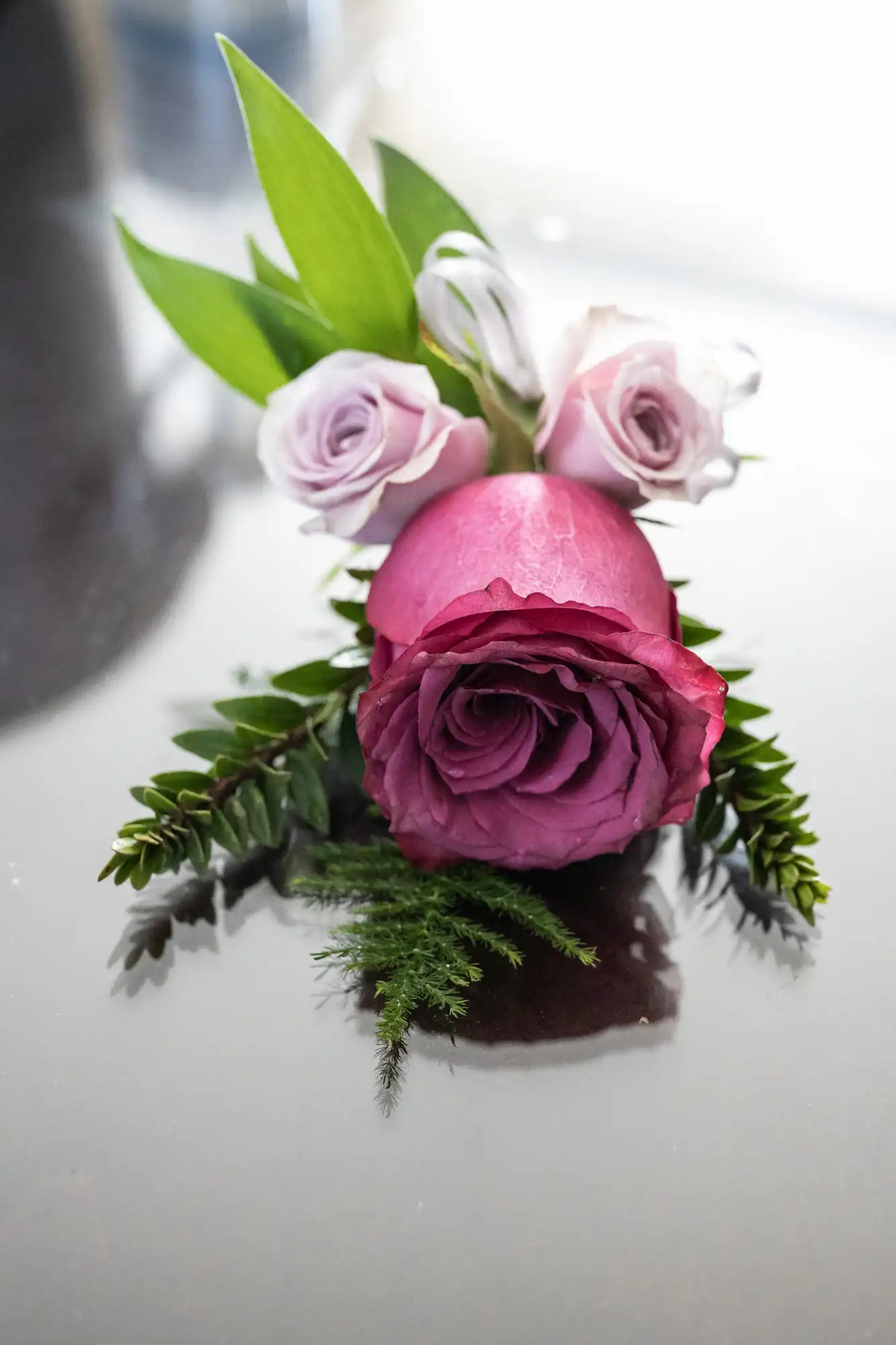 Close-up of a floral arrangement featuring one large and three small roses in varying shades of pink and purple, complemented by green leaves, placed on a reflective surface.