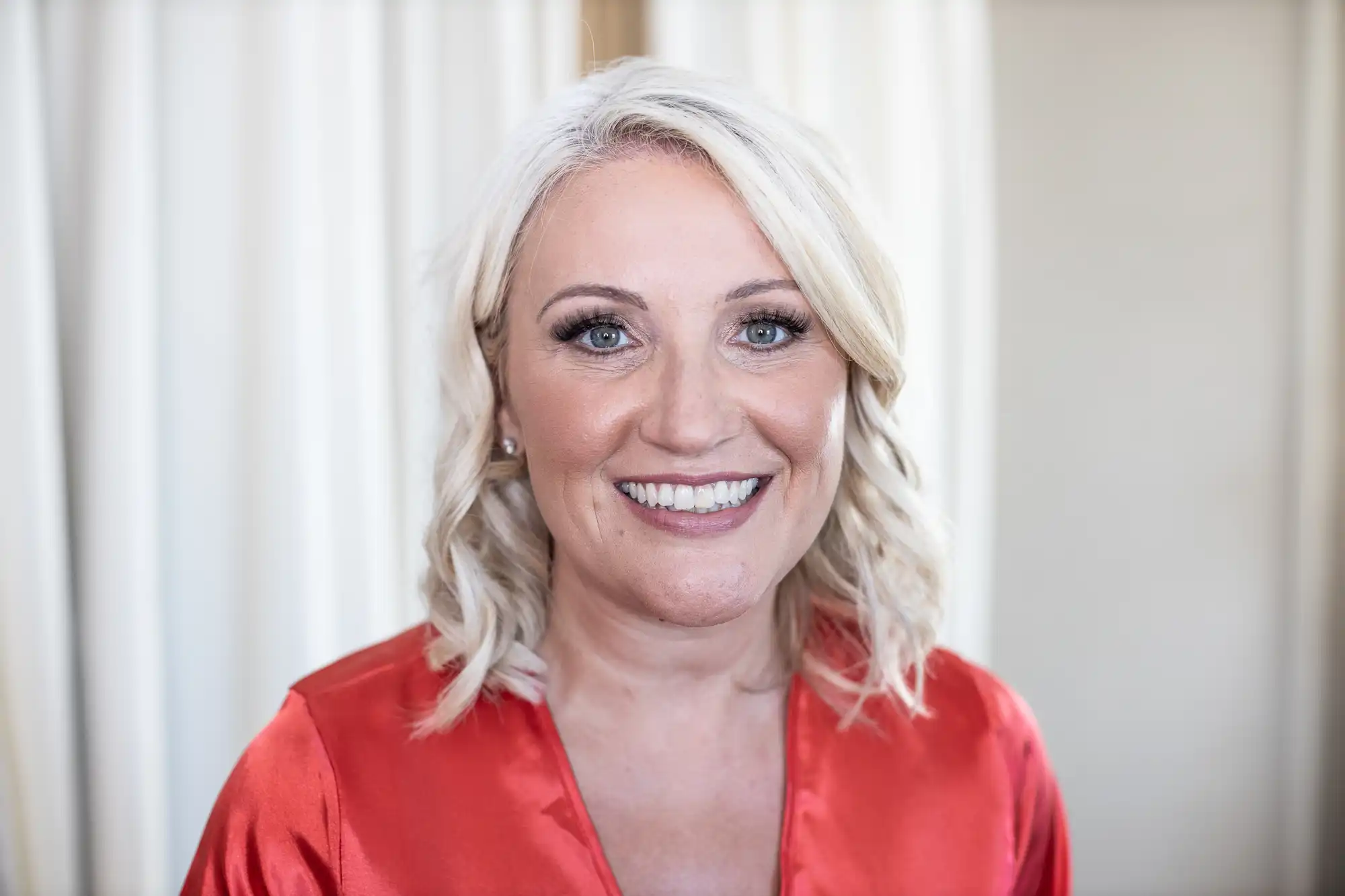 A woman with blonde hair smiles at the camera, wearing a red V-neck top, standing in front of light-colored curtains.