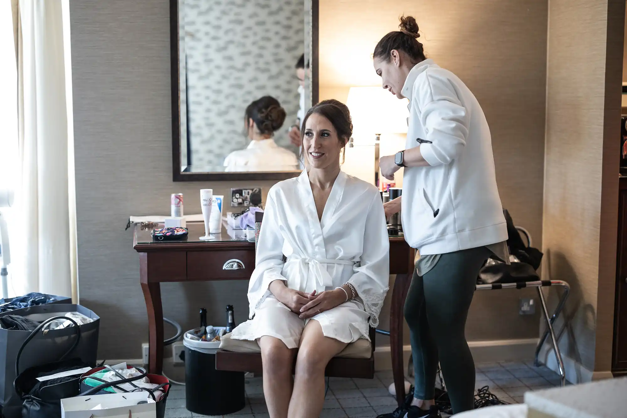 A woman in a white robe sits at a vanity table while another person styles her hair in a room with various beauty products and a lamp.