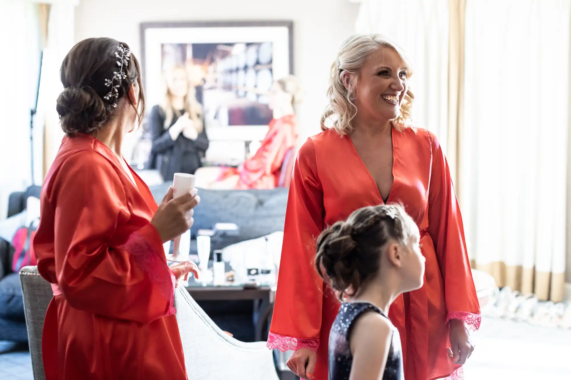 Three women in red robes and a young girl with an updo hairstyle stand in a bright room, smiling and chatting. Other people are blurred in the background.