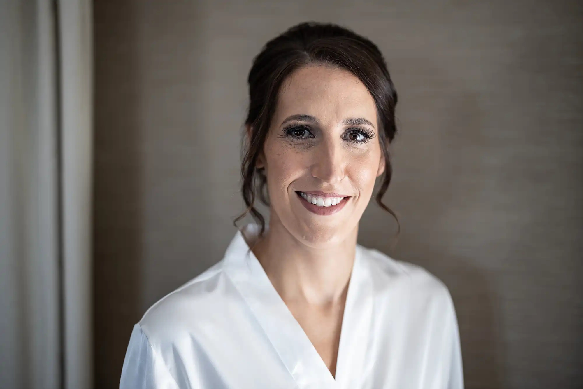 A woman with dark hair styled in an updo and wearing a white robe smiles while looking at the camera in a softly lit indoor setting.