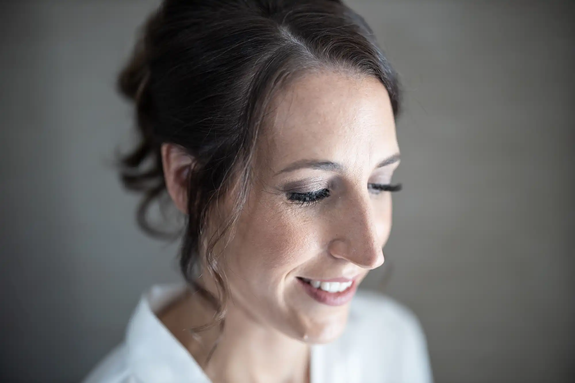 A close-up of a woman with brown hair, styled in a loose updo. She is wearing a white top and smiling with her eyes closed. The background is blurred and gray.