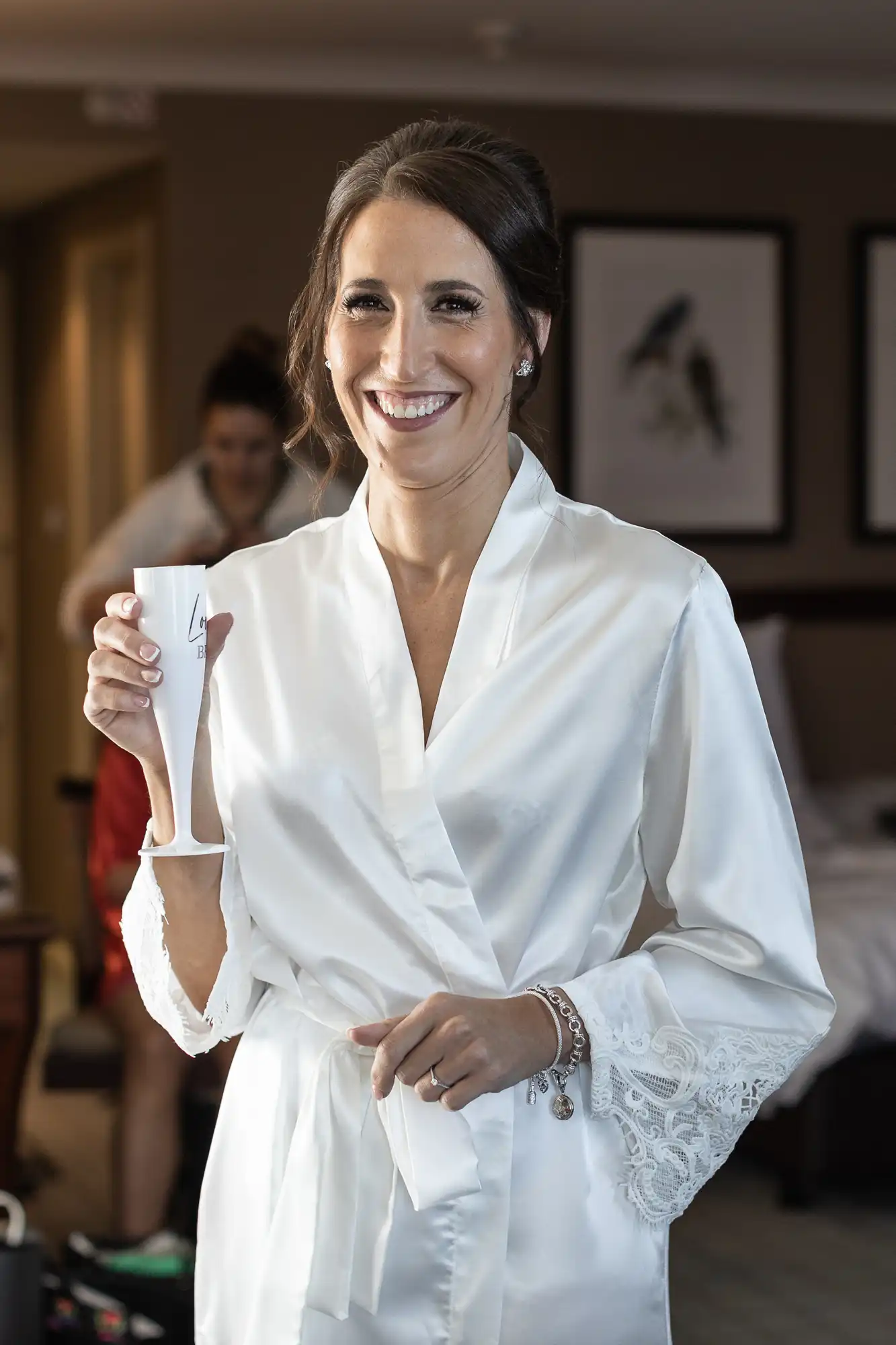 A woman in a white robe smiles and holds a small folded piece of paper in a hotel room. Another person is in the background.