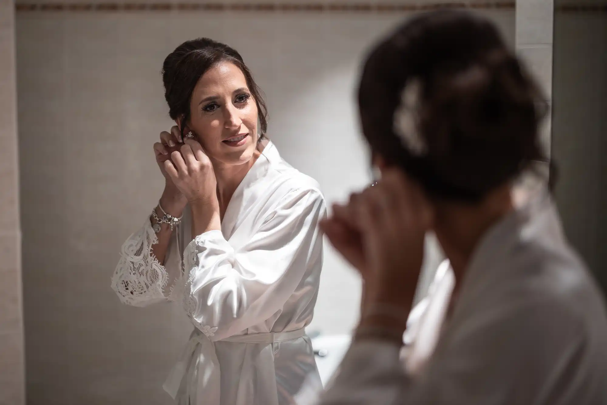 A woman in a white robe stands in front of a mirror, adjusting an earring while her reflection is visible.