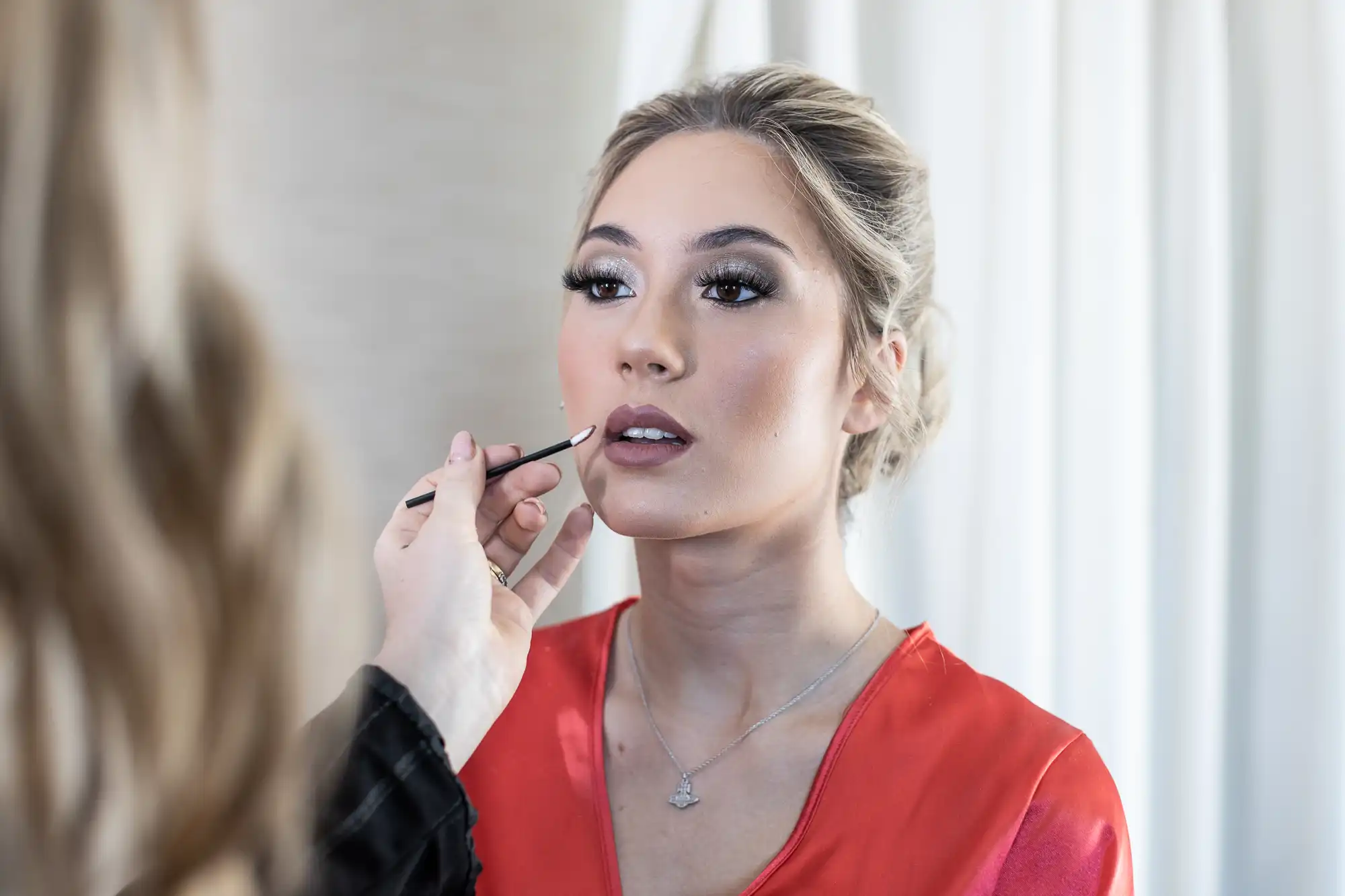 A woman in a red top has makeup applied to her lips by another person.