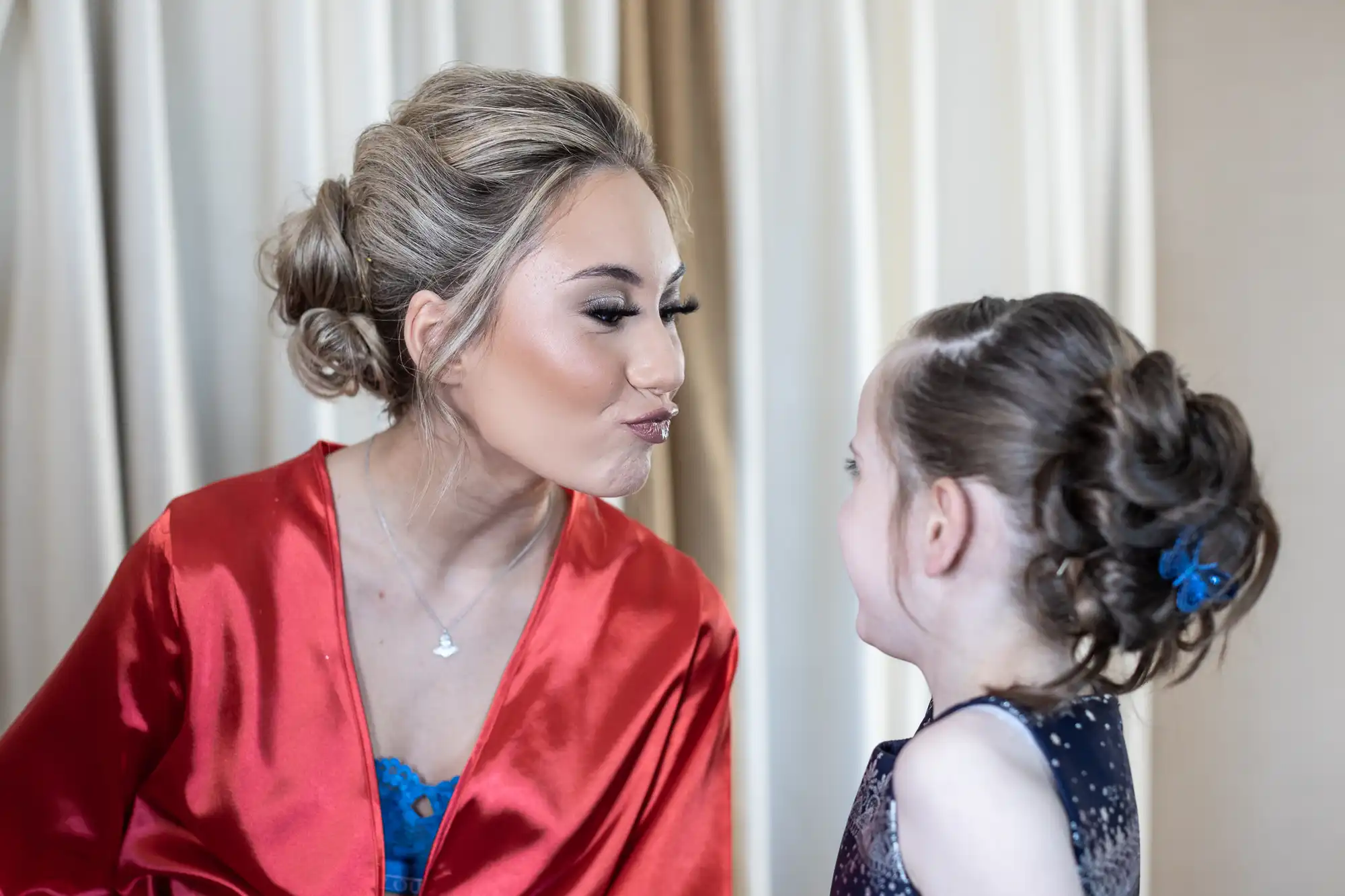 An adult in a red robe playfully puckers lips at a little girl with an updo hairstyle, both smiling in a bright indoor setting.