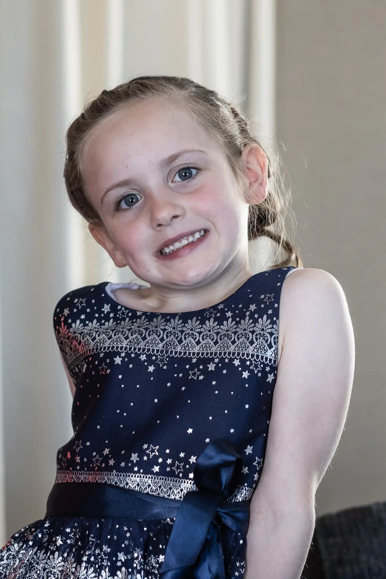 Young girl with light skin and braided hair, smiling and wearing a blue dress with white patterns and a ribbon.