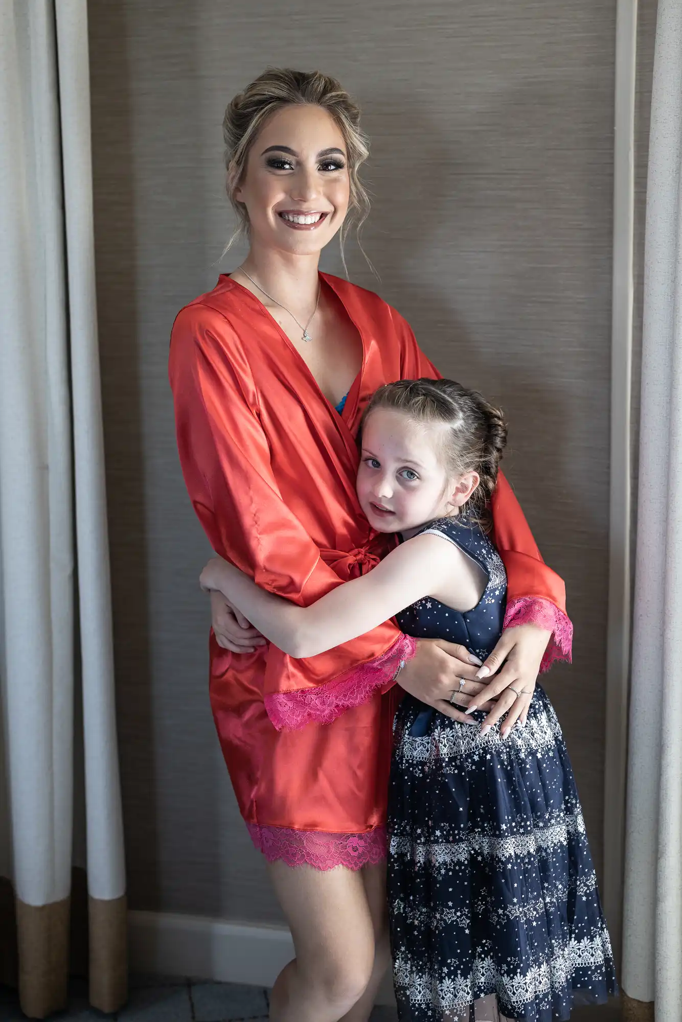 An adult in a red robe with pink trim hugs a young child in a blue dress with white patterns. Both are smiling and standing indoors.