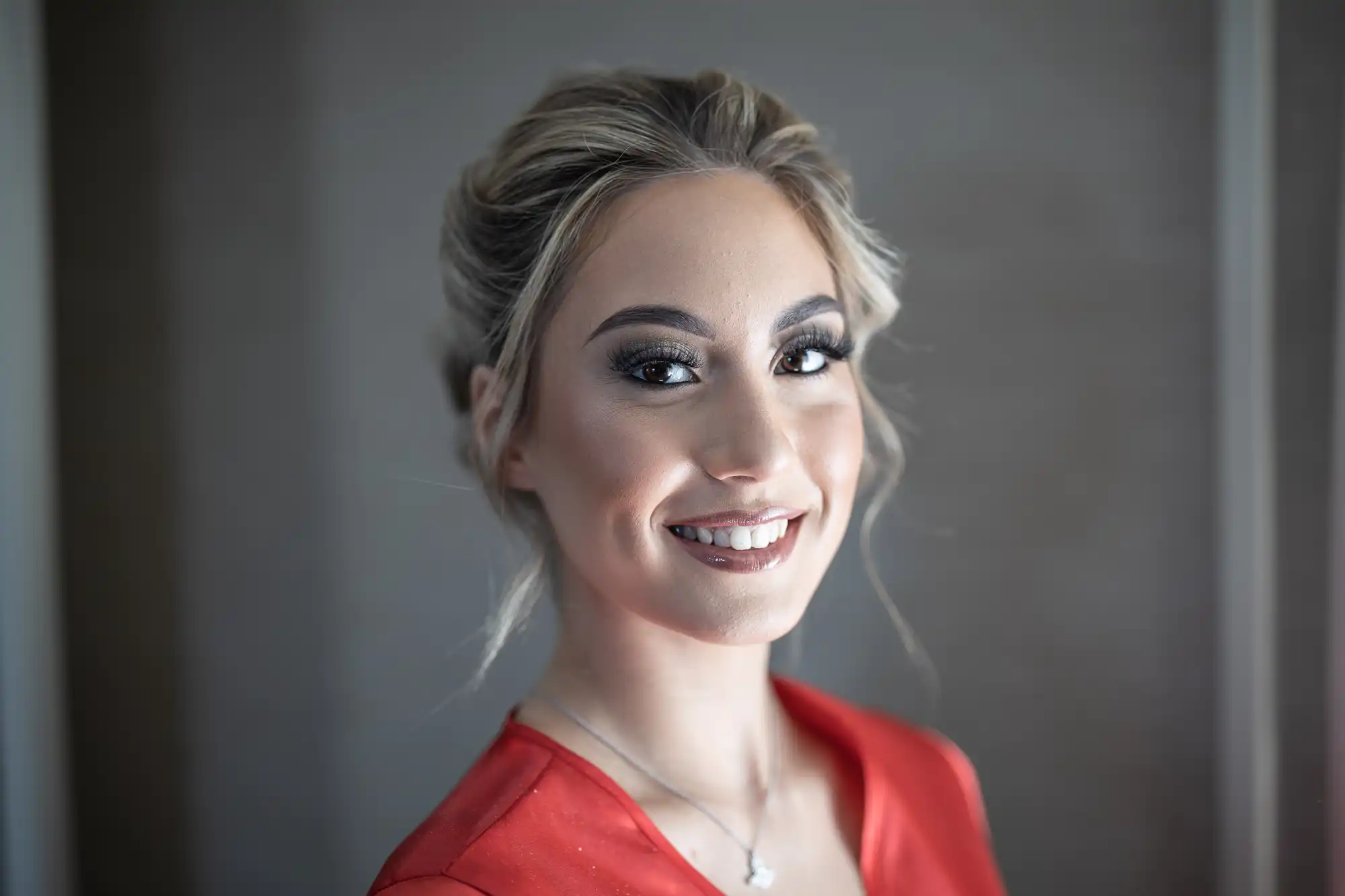 A woman with styled hair and makeup, wearing a red top and a necklace, smiles at the camera.