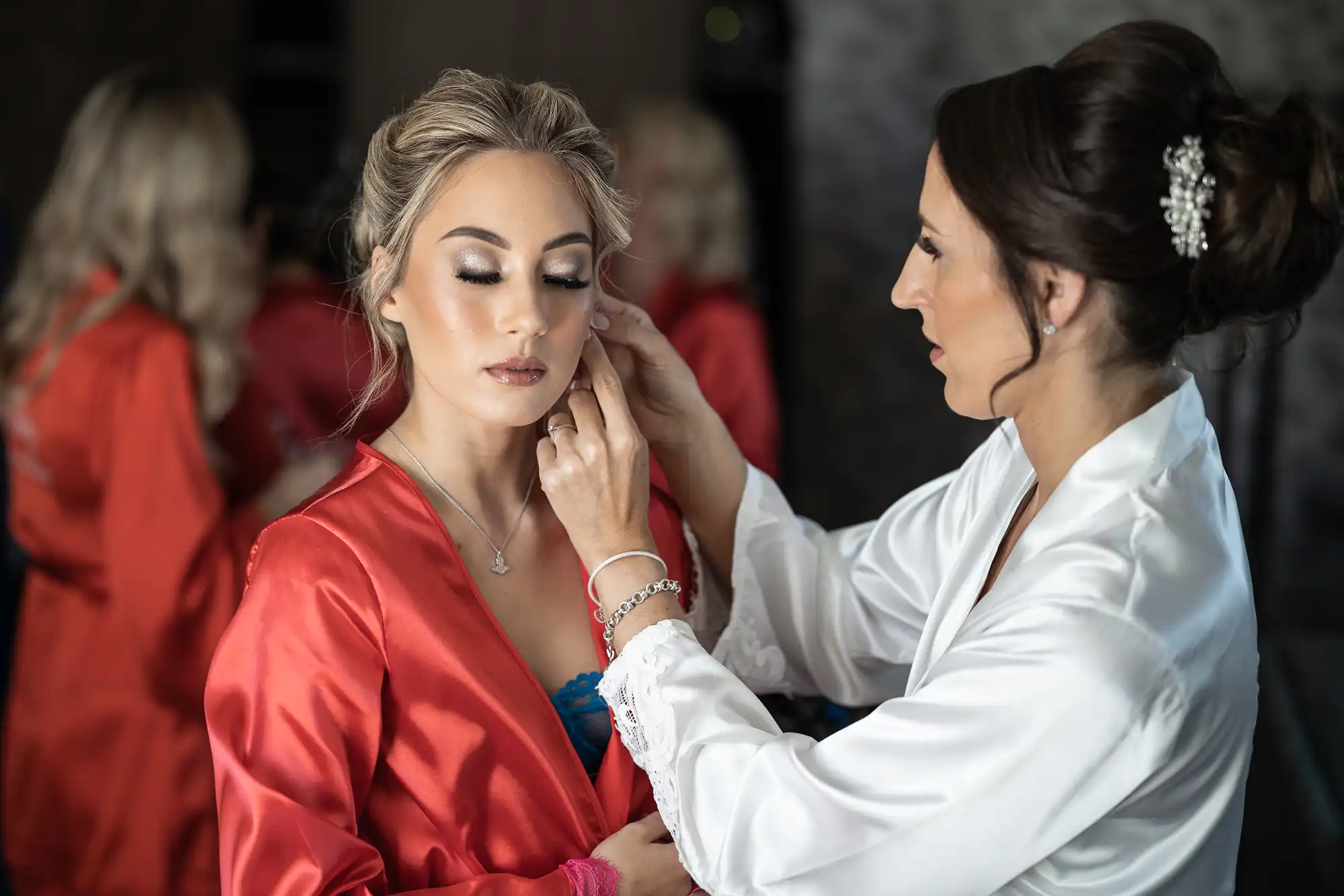 A woman in a white robe helps another woman in a red robe prepare by adjusting her earring.