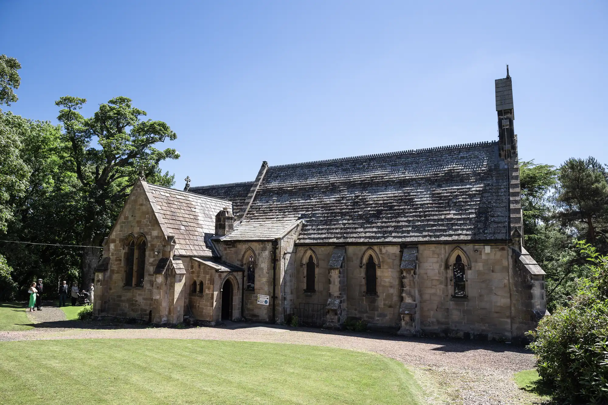 A historic stone church with arched windows and a pitched roof, surrounded by trees and grass, under a clear blue sky.