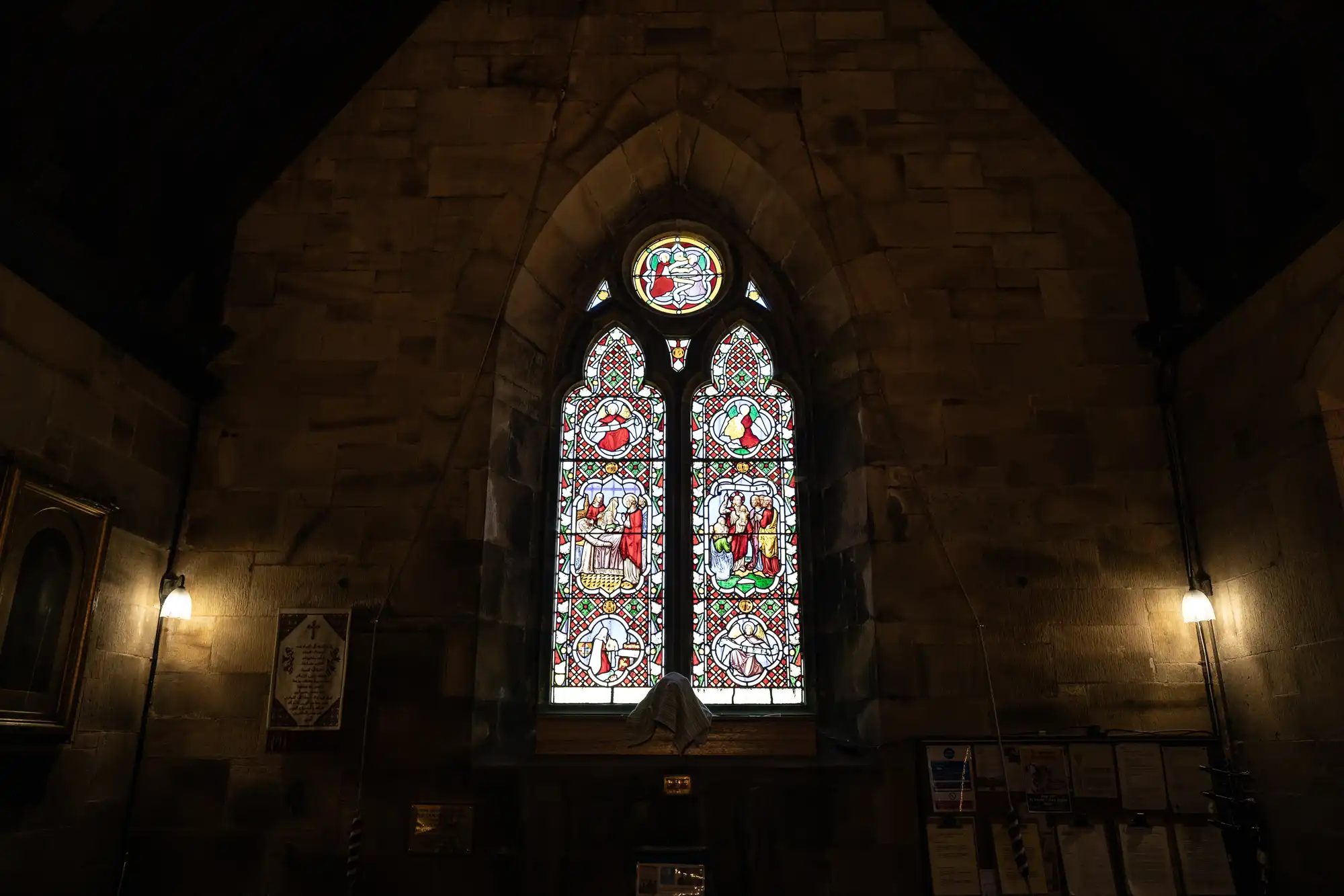 A stained glass window with intricate designs in a dimly lit stone building, featuring religious motifs and a pointed arch. The surroundings include some framed items and a softly illuminated interior.