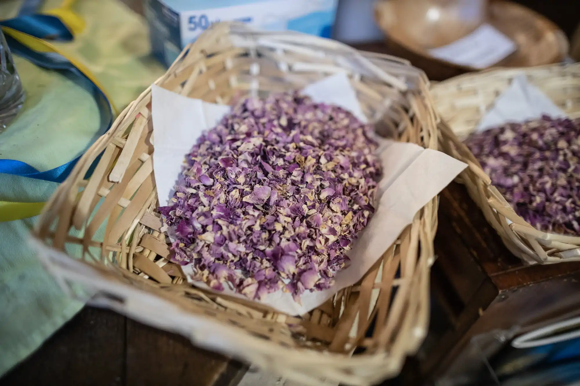 A wicker basket filled with dried purple flowers rests on a wooden surface, lined with white paper. Additional items are partially visible in the background.