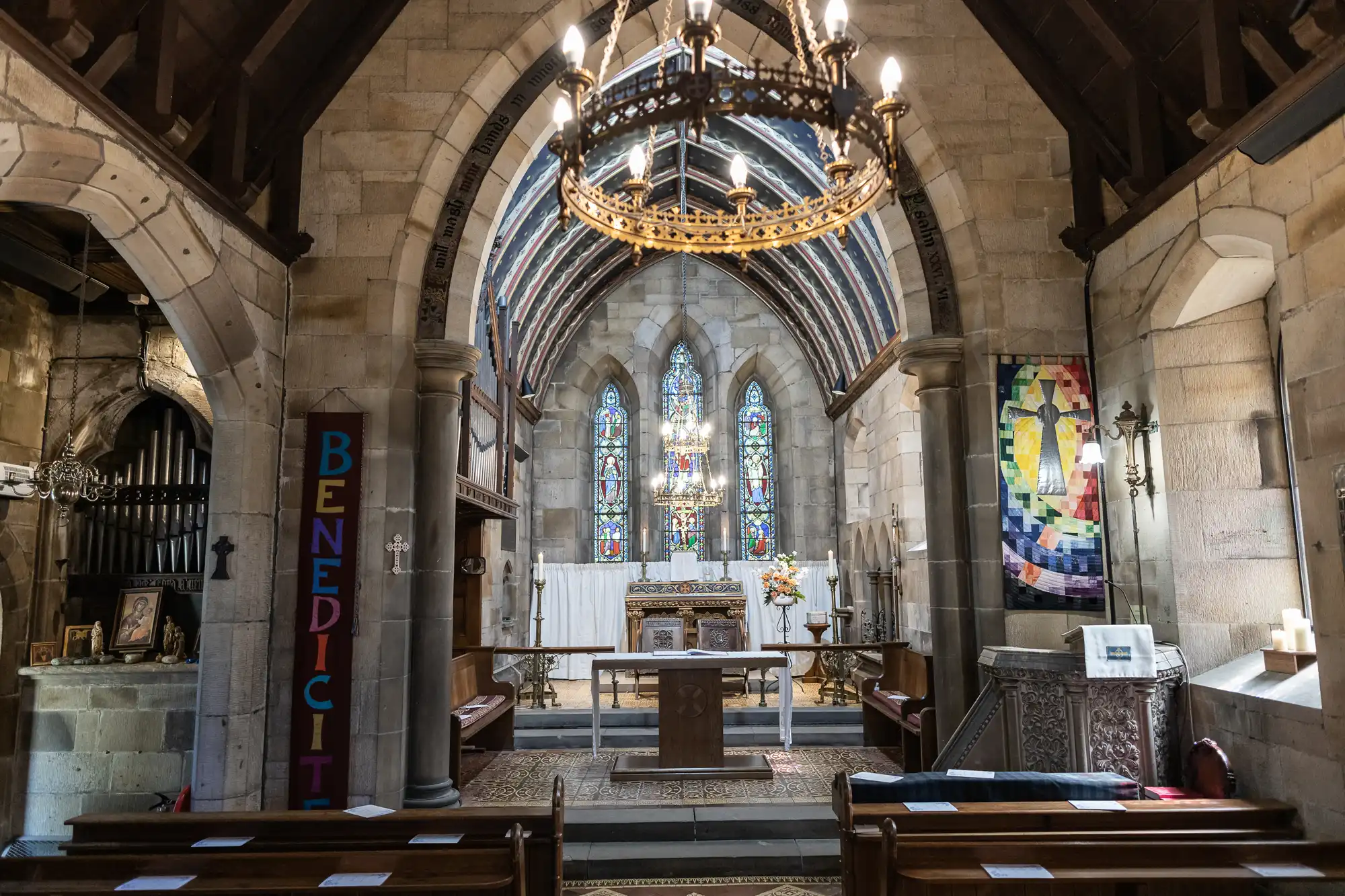 Interior of a church with stone walls and arched ceilings. A chandelier hangs above the aisle. The altar is adorned with flowers, and stained glass windows are visible in the background.